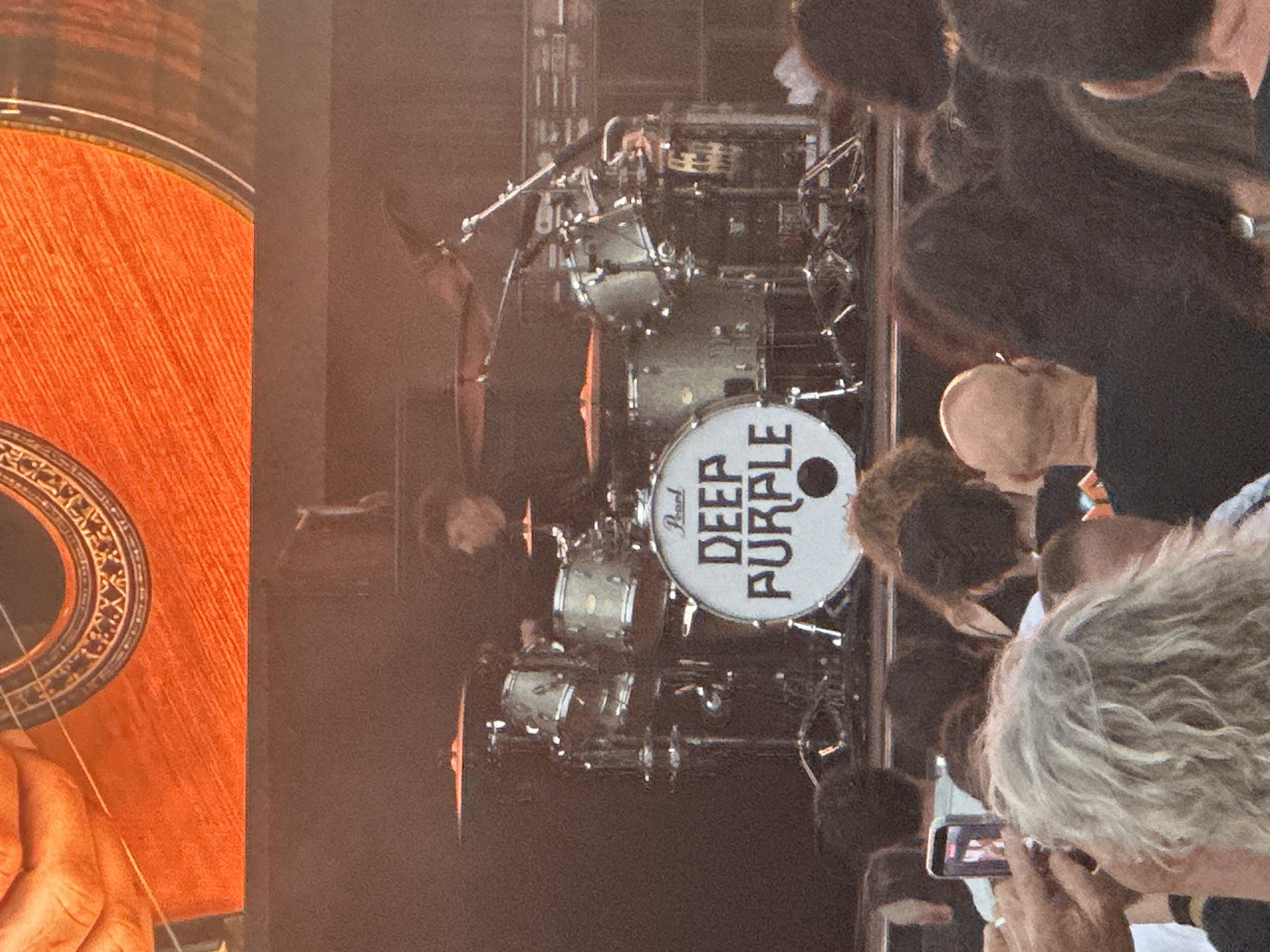 This image captures a live music performance at an outdoor venue. In the foreground, a crowd of people is visible, with some individuals holding up smartphones to record the event. The background features a drummer on stage, seated behind a drum kit emblazoned with the "Deep Purple" logo. The stage is lit, creating a dramatic effect. The overall scene suggests a concert atmosphere, likely in a city like Jerez De La Frontera, Spain, known for its vibrant cultural events.