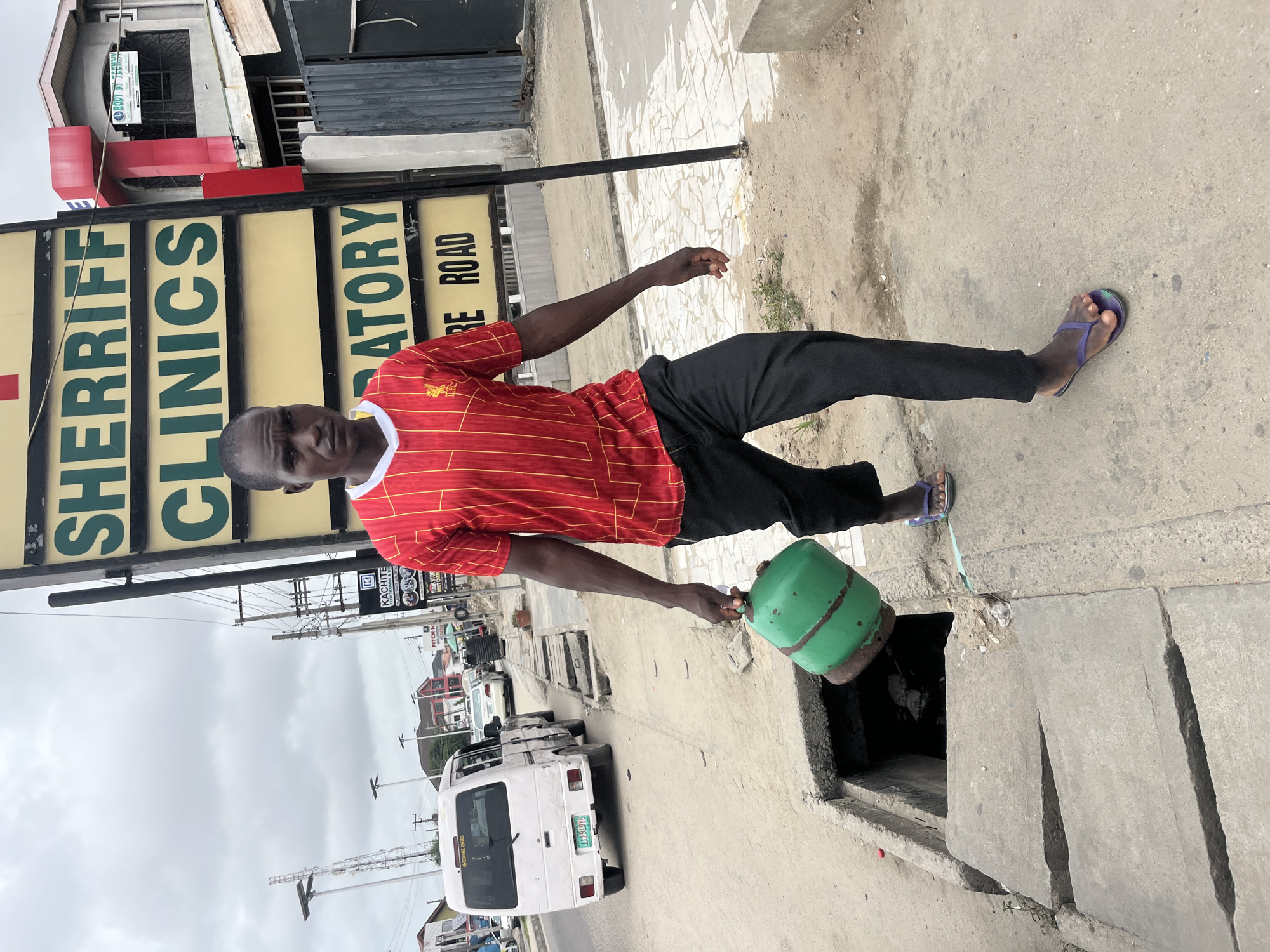 A man stands on a sidewalk in Ado, Nigeria. He wears a red shirt with gold stripes, black pants, and purple sandals. He holds a green, cylindrical object over an open sewer grate in the sidewalk. Behind him is a street with vehicles, including a white van. A sign reading "Sheriff Clinics Laboratory" is visible in the background. The sky is overcast.