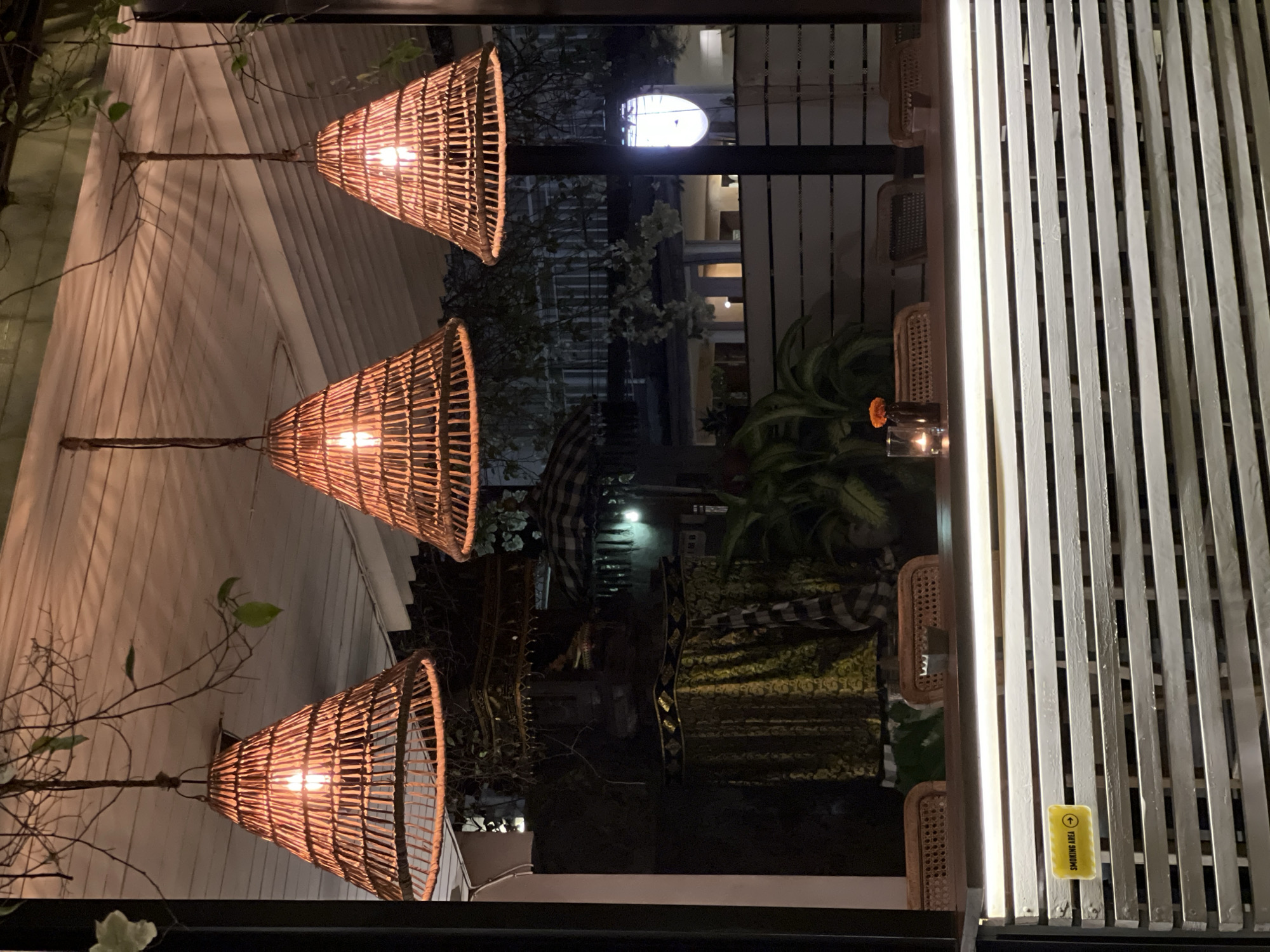The image depicts an outdoor scene in North Kuta, Indonesia. Three wicker lamps hang from the ceiling. The scene appears to be a restaurant patio at night. Tables are visible with rattan chairs. A "smoking area" sign is affixed to a partition consisting of vertical white planks. Decorations including flowers and patterned fabric are also observed.