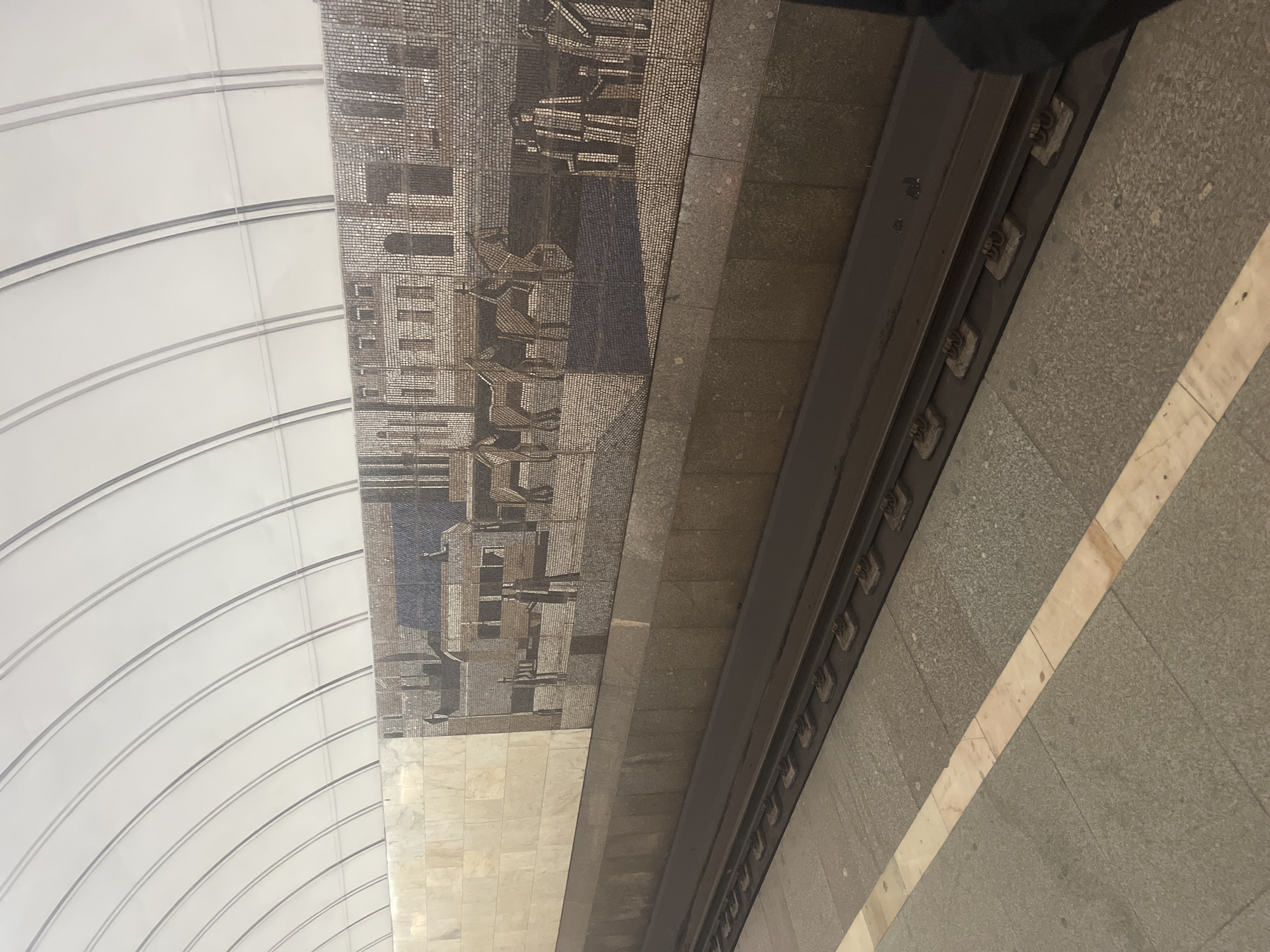 The image shows a mosaic on the wall of a metro station in Moscow, Russia. The mosaic depicts a historical scene with buildings, people, and horses. The station has a vaulted ceiling with white panels and metal supports. The tracks of the metro line are visible in the foreground, with a platform next to them. The mosaic appears to be made of small, colorful tiles and is a significant piece of public art. The overall impression is one of historical significance and artistic merit within a functional public space.