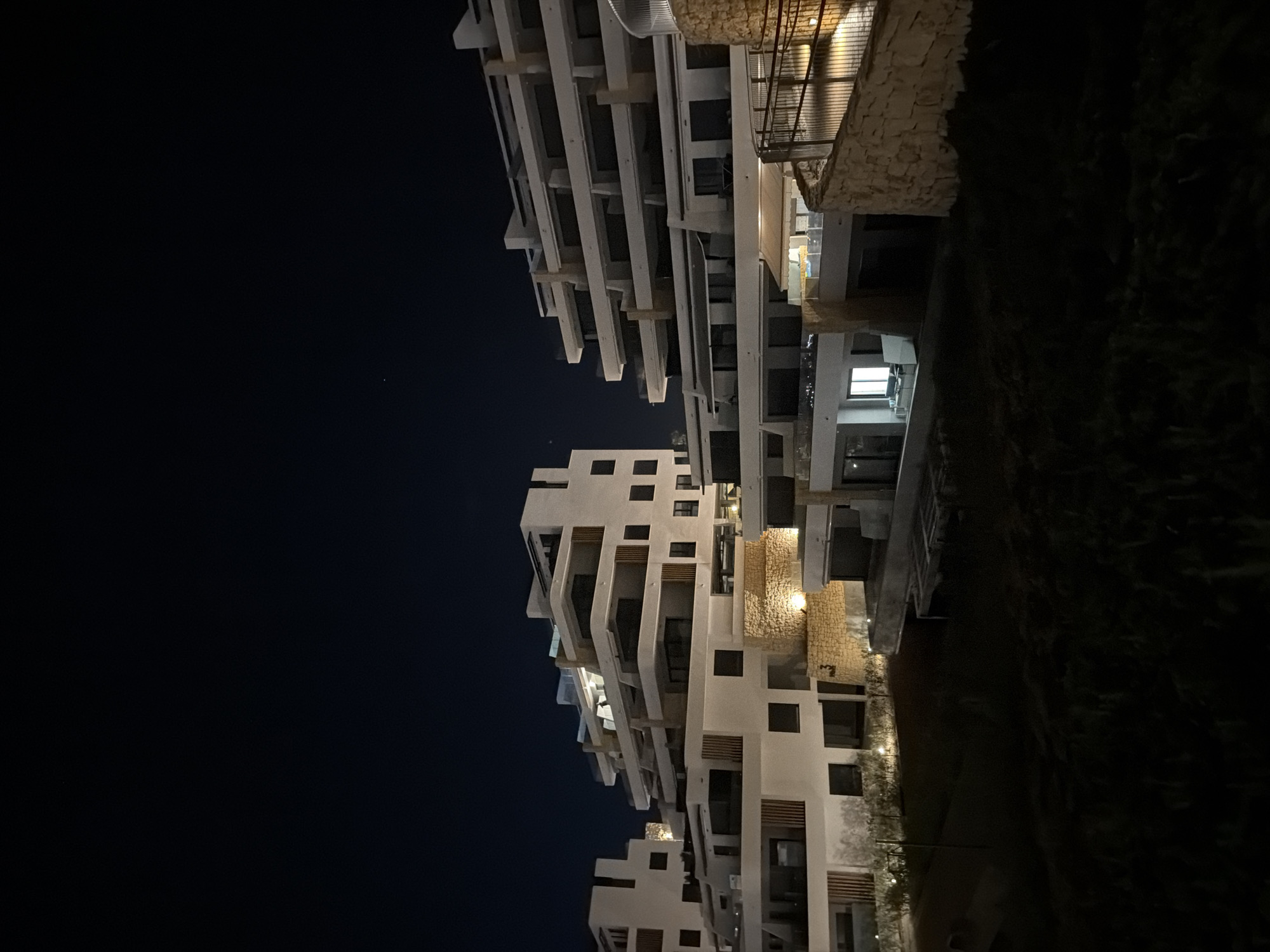 This is a nighttime exterior, low-angle shot of a modern, multi-level apartment complex in Finestrat, Spain. The buildings are constructed from white concrete and feature numerous balconies, large dark-tinted windows, and light-colored wooden accents. The architecture is characterized by terraced levels that step down the hillside, creating a tiered effect. Stone retaining walls, illuminated by warm-toned lights, are visible in the foreground and between building levels, adding texture and contrast. The sky above is dark blue, indicating late evening or night. The foreground is dominated by dark, shadowy landscaping and grass. No people or vehicles are visible in the image. The scene suggests a residential or resort property, with some lights on within the apartments, indicating occupancy.