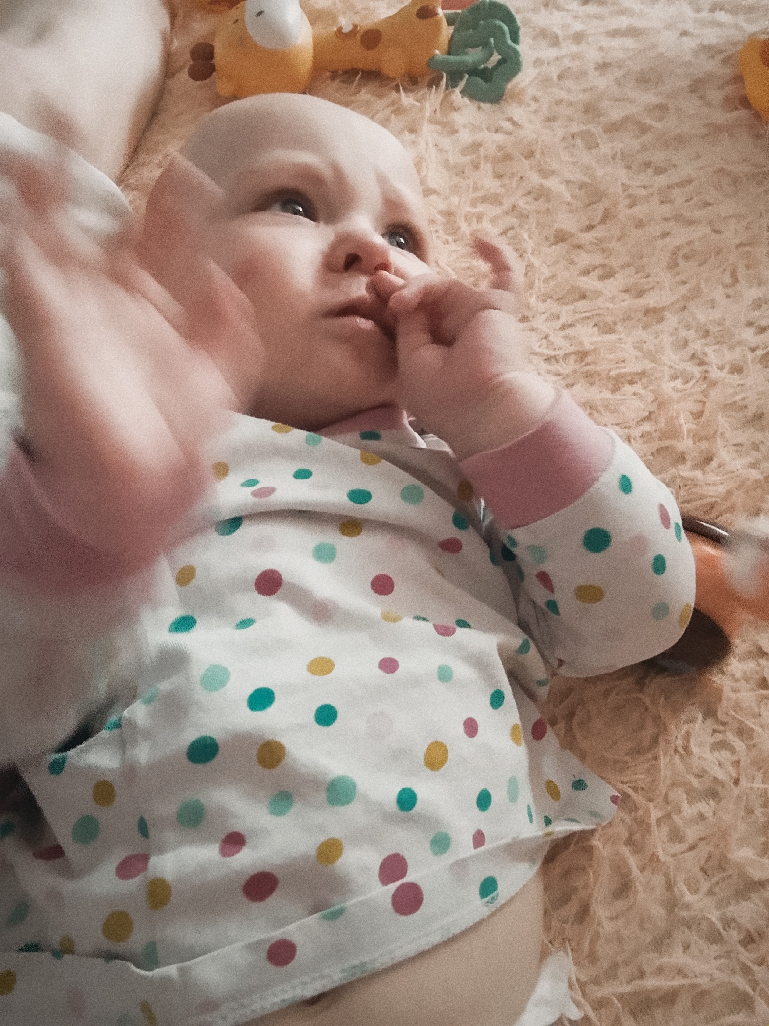 An overhead, medium shot captures a bald infant lying on a textured, beige surface, likely a rug or blanket. The baby is wearing a white onesie adorned with multi-colored polka dots in pink, yellow, and teal. The infant's right hand is raised, with fingers blurred due to motion, and appears to be touching its face, near the nose. The baby's head is turned slightly to the left, with its gaze directed upwards and to the left. In the background, out of focus, are several baby toys, including a yellow giraffe-like toy and a green toy. The overall lighting is warm and soft, suggesting an indoor setting. The image does not provide any information that would allow for the determination of a specific location in Minsk, Belarus.