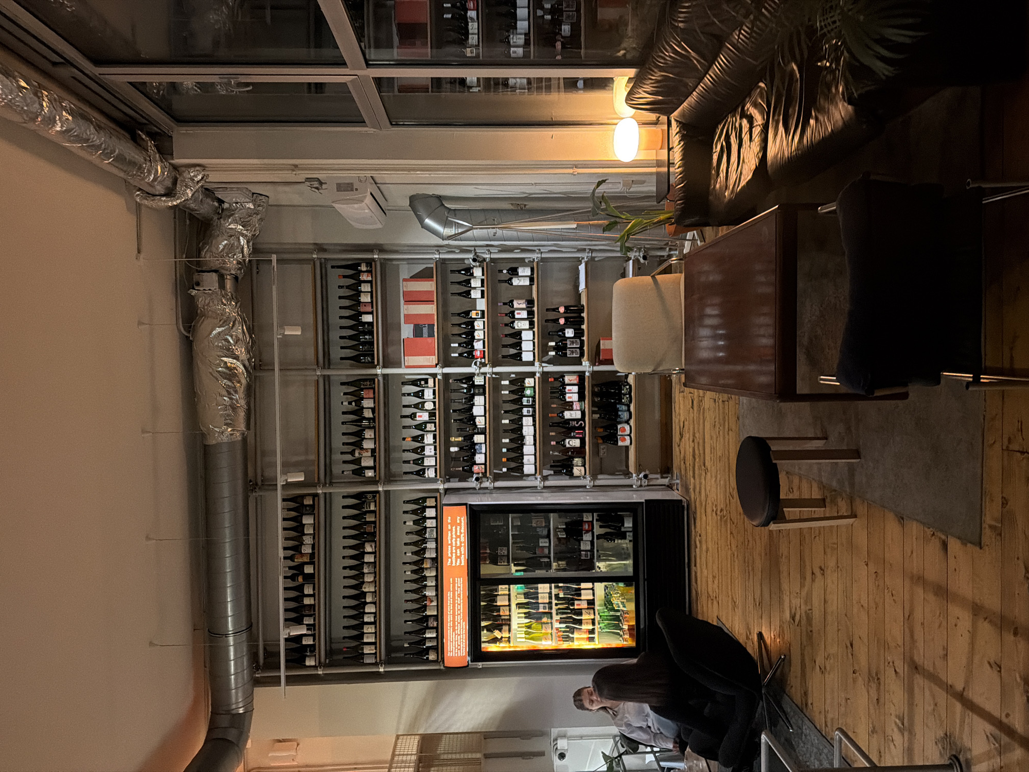 The image displays the interior of a commercial establishment, specifically a wine bar or shop, located in Belgrade, Serbia. The scene is illuminated with warm, dim lighting.
The dominant feature is an extensive, floor-to-ceiling metal shelving system along the left wall, densely stocked with hundreds of wine bottles arranged in horizontal rows. Centrally positioned within this shelving system is a black, two-door refrigerated display unit, internally illuminated, also containing numerous bottles. Above this unit, an orange sign with white text is visible.
The flooring consists of light-colored wooden planks, partially covered by a dark rectangular rug in the central area. Seating arrangements include a dark brown, distressed leather sofa positioned along the right side of the frame, accompanied by a rectangular dark wooden coffee table in front of it. A light-colored armchair is partially visible behind the table. A small, dark-topped stool with light wooden legs is placed on the rug in the mid-ground. In the left foreground, a black lounge chair with a prominent base is occupied by an individual with dark hair, seen from behind. To their immediate left, another individual with light brown hair is seated on a light-colored armchair or sofa, with their profile partially visible.
The ceiling features exposed metal air ducts, some wrapped in silver insulation. Large, multi-pane windows are visible on the far right wall, and an air conditioning unit is mounted below them. A small potted plant rests on a low table between the leather sofa and the right wall, next to a white mushroom-shaped table lamp emitting a warm glow.