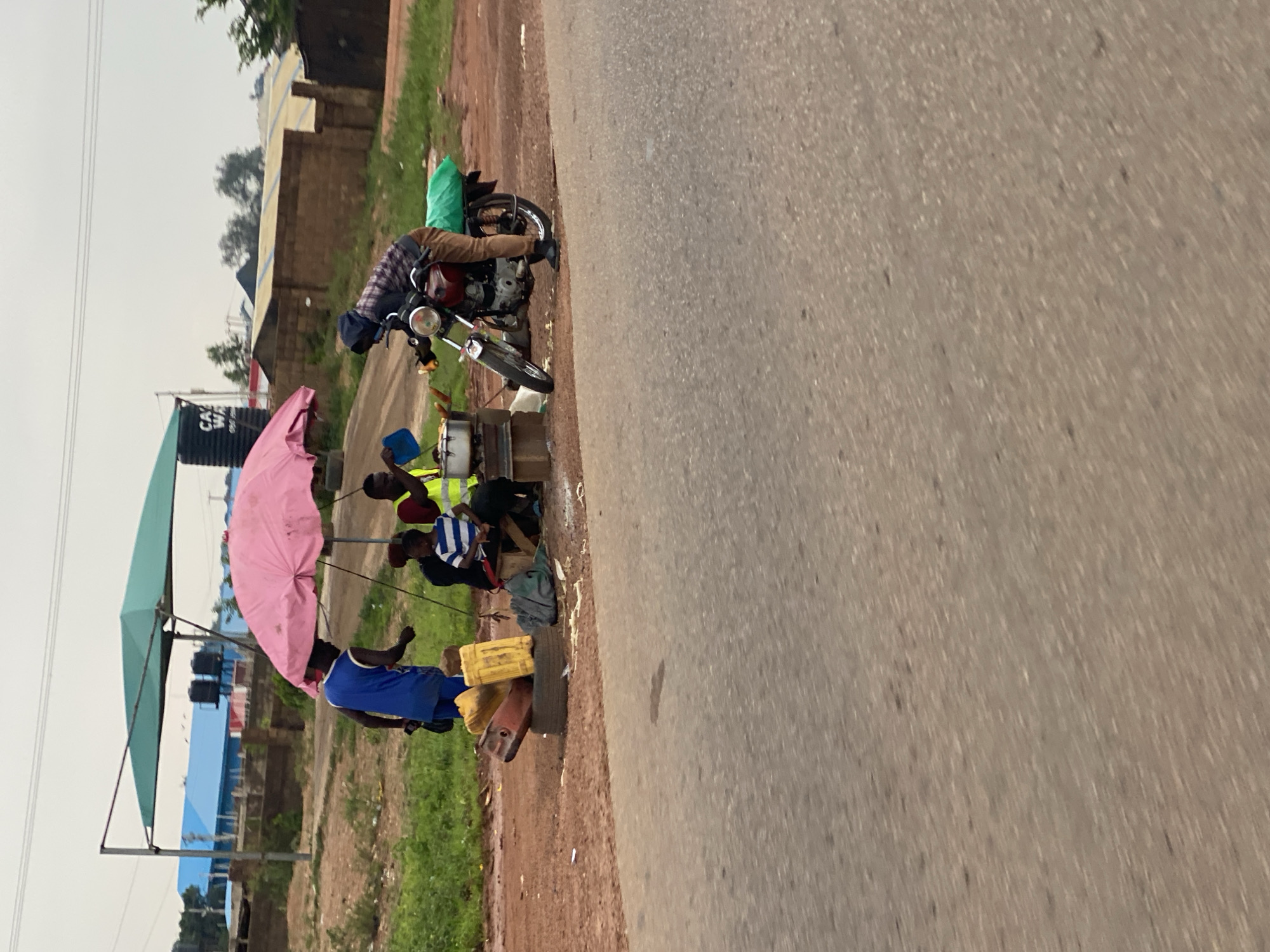 A street-side scene in Jos, Nigeria, features several individuals engaged in commerce and transportation. Under a large, pale pink umbrella, a man in a blue tank top and shorts stands near a collection of yellow jerrycans and a black tire. To his right, two other individuals are seated, one in a blue and white striped shirt and the other in a yellow vest, appearing to interact with cooking equipment. A motorcycle, with a green package secured to its rear, is parked beside a person in a hooded jacket and brown pants, who is looking towards the cooking area. In the background, buildings with blue roofs and a water tank with "CA WASH" visible on it are present, along with a dirt road and lush green vegetation. The foreground is dominated by a grey asphalt road.