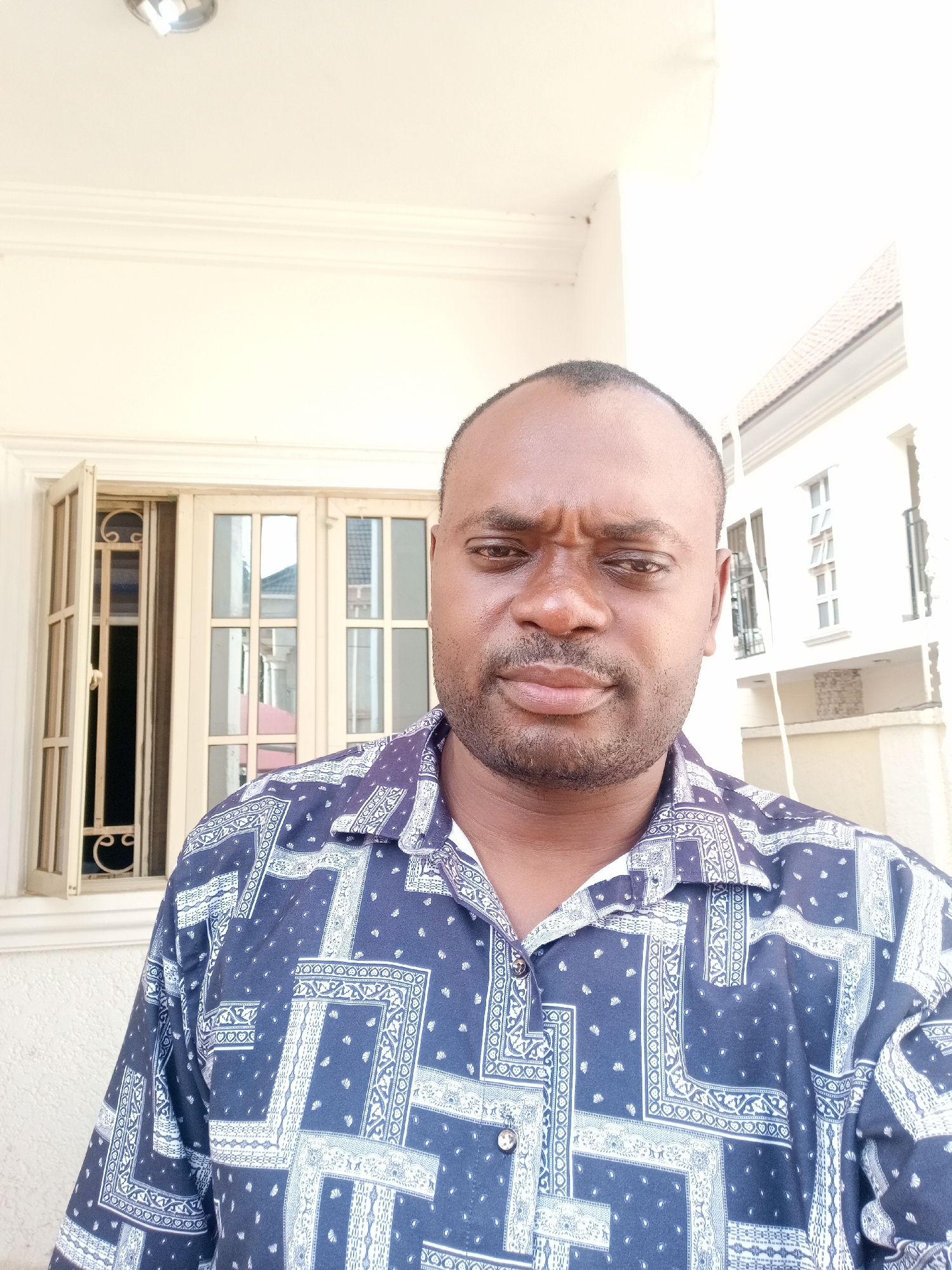 A medium-close shot captures a middle-aged man with dark skin, a partially shaved head, and a well-groomed beard. He is wearing a blue patterned shirt with a white collar. The man is positioned in front of a building exterior, with an open window to the left and a partial view of another building behind him on the right. The background structures are painted in light, neutral tones. The man's facial expression suggests a stern or critical demeanor. The location is identified as Gwarinpa, Nigeria.