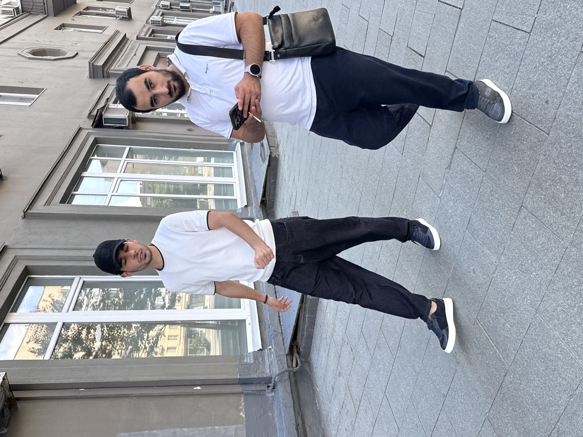 Two men are walking on a paved sidewalk in front of a building. The man on the left is wearing a white t-shirt, black pants, and a black baseball cap. The man on the right is wearing a white polo shirt, dark blue pants, and carrying a black messenger bag over his shoulder. He is holding a smartphone in his left hand and has a smartwatch on his right wrist. The building behind them is light gray with multiple windows and air conditioning units visible. The sidewalk is made of gray rectangular paving stones. The environment suggests an urban setting, likely in Moscow, Russia, given the architectural style and urban elements.