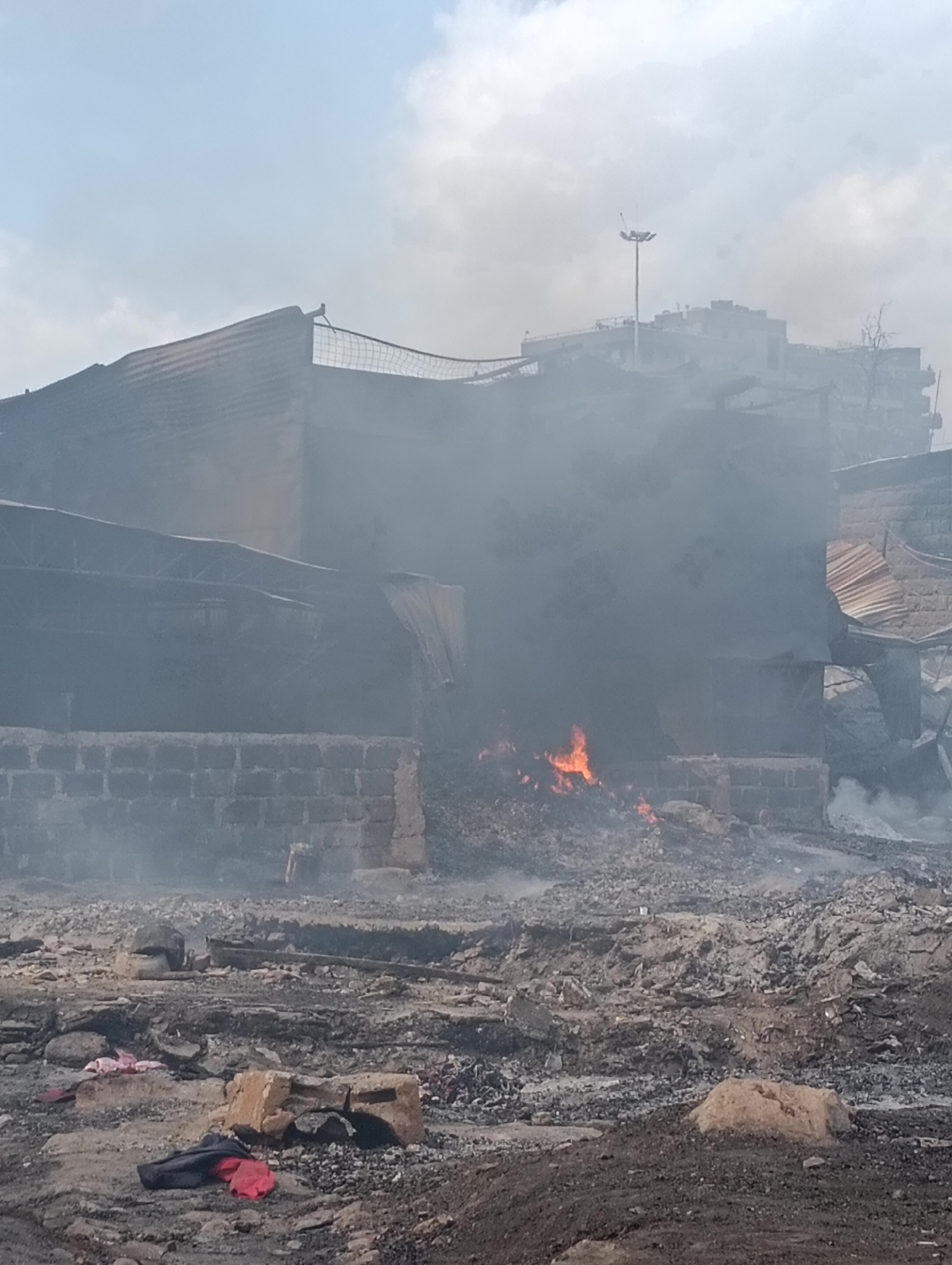The image depicts the aftermath of a fire in a densely populated urban area, believed to be Nairobi, Kenya. A building, partially constructed of brick, is engulfed in thick, dark smoke. Flames are visible at the base of the structure, indicating ongoing combustion. Debris, including rubble and charred materials, litters the foreground and surrounding area, suggesting significant destruction. In the background, taller buildings are partially obscured by the smoke, with a tall light pole visible above one of them. The sky is overcast with clouds. Scattered on the ground in the foreground are pieces of fabric, possibly clothing, among the rubble. There are no people clearly visible in the image, though the presence of clothing suggests recent human activity. The scene is chaotic and indicative of a significant fire incident.