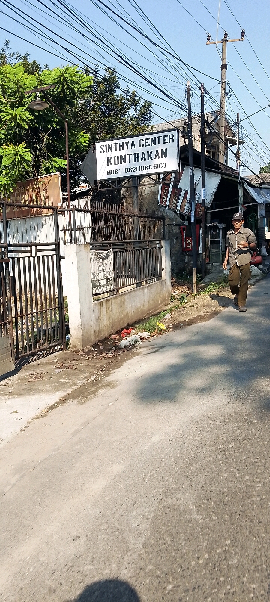 This image shows a street scene in Depok, Indonesia. A man in a light brown shirt and dark pants is walking on the right side of the road. He appears to be holding a phone or similar device. A sign that reads "SINTHYA CENTER KONTRAKAN HUB: 08211088 6163" is visible on the left side of the road, above a metal fence and a concrete wall. The area is lined with trees and electrical wires. There is some debris and trash on the side of the road. The sky is clear and blue. The scene appears to be in a residential or commercial area.