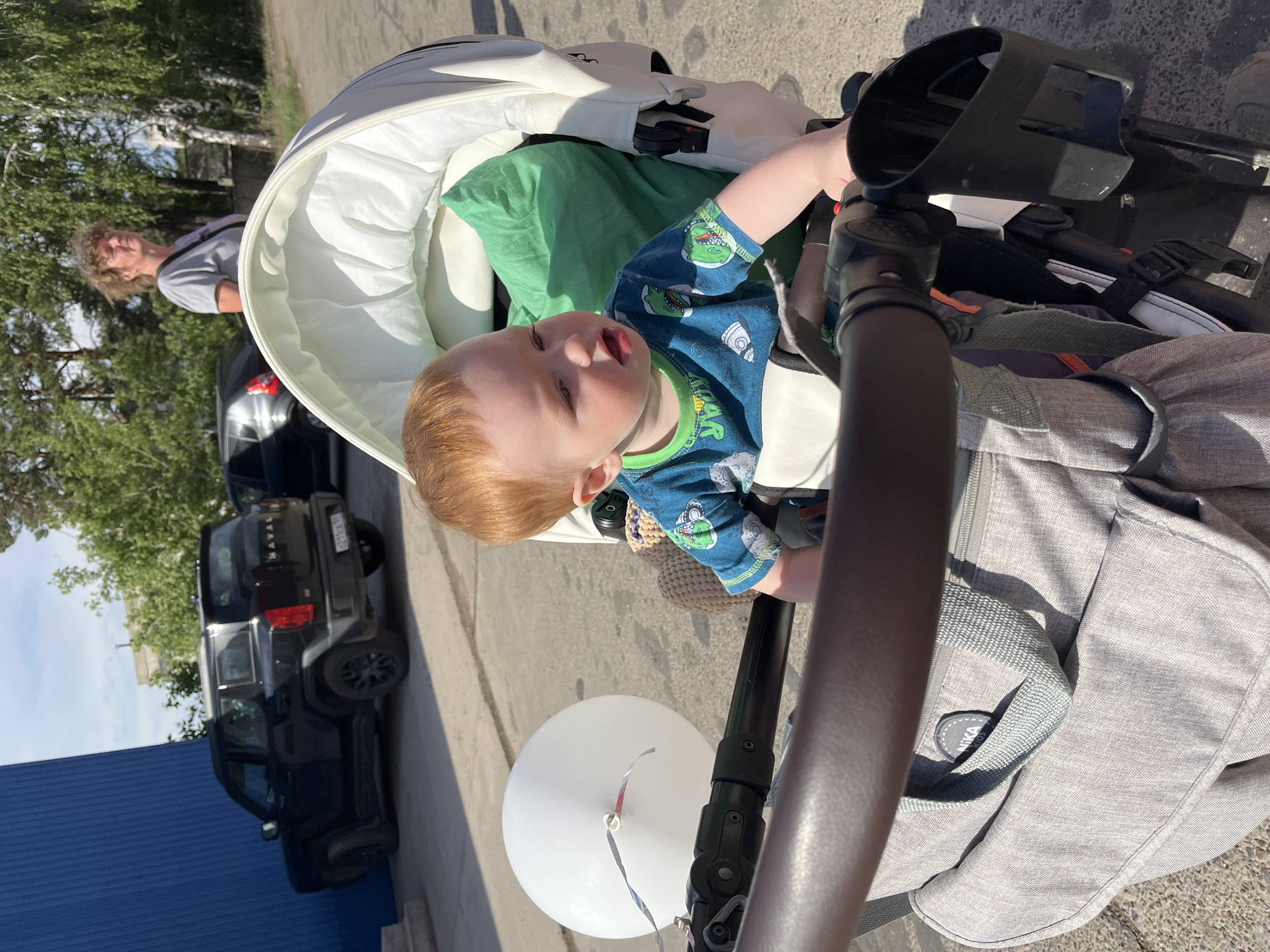 A young child with red hair is sitting in a white stroller with a green blanket inside. The child is wearing a blue shirt with dinosaur print. A black SUV is parked on the left side of the frame. A man with long hair is standing in the background. The stroller has a gray bag attached to it, with a white balloon tied to the handle. The scene appears to be outdoors on a sunny day, likely on a paved surface, with a blue building visible in the lower-left corner. This scene takes place in the city of Chaikovsky, Russia.