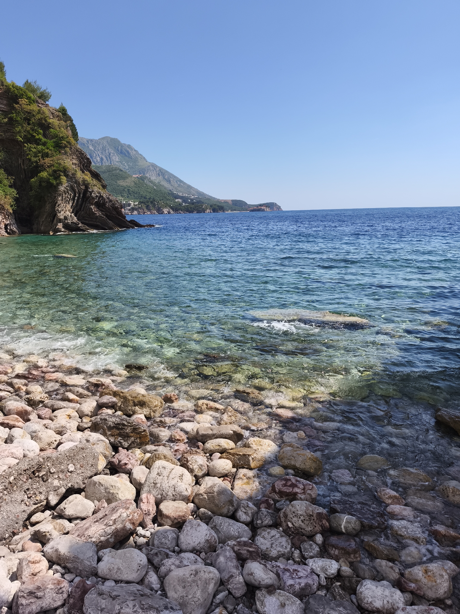 The image depicts a rocky beach on a sunny day in Rafailovići, Montenegro. In the foreground are numerous grey, white, and reddish-brown rocks of varying sizes, partially submerged in clear, turquoise water. The water clarity allows visibility of the underwater rocks and seabed. In the midground, the water deepens to a darker blue. Further out, the horizon meets a clear, light blue sky.
To the left, a cliff face covered in green vegetation rises abruptly from the shoreline. In the distance, a mountain range and a settlement with buildings can be seen along the coast. The overall impression is one of a natural, coastal landscape in good weather.