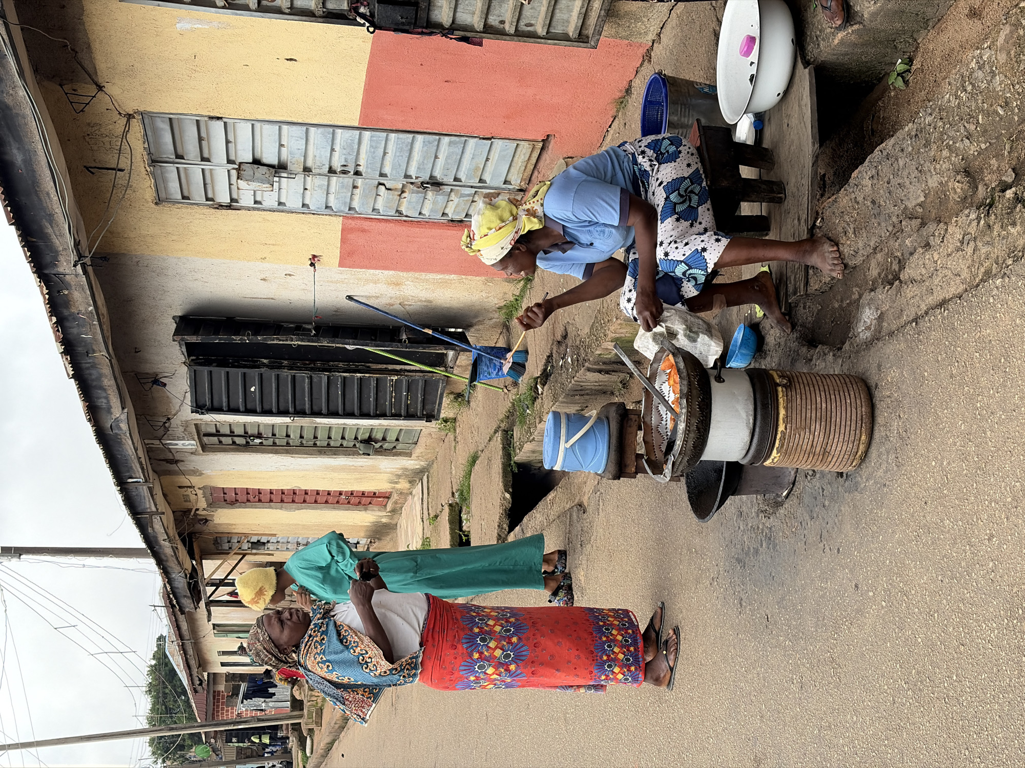 In Jos, Nigeria, two women are observed in conversation on a dusty street. One woman, adorned in a vibrant red and blue patterned skirt and a patterned headscarf, stands to the left. She is engaged in conversation with another woman, who is dressed in a teal garment and a yellow headscarf. To the right, a woman is seated on a small wooden stool, wearing a blue shirt and a blue and white patterned skirt. She holds a stick in her hand, appearing to be cooking or tending to food in a large metal pot placed on top of a stacked cooking apparatus. Several containers and bowls are visible around her, suggesting a street food vending setup. The backdrop consists of buildings with textured walls painted in yellow and red, featuring metal shutters on the doors. A drainage ditch runs along the side of the street, and overhead power lines are prominent. The scene depicts a typical street-side activity, likely a marketplace or a residential area with commerce.