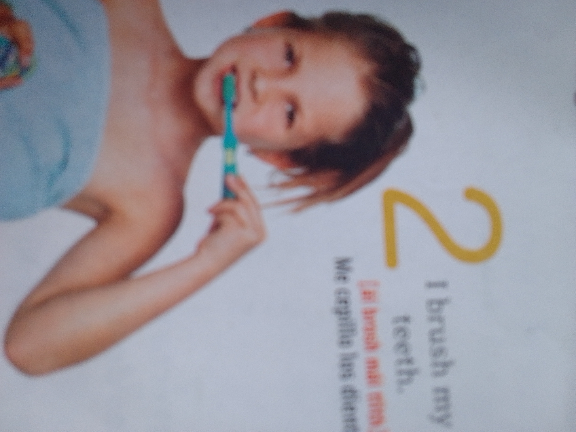 The image shows a young person, likely a child, with brown hair and fair skin, holding a blue toothbrush. The child is positioned in a way that their upper body is visible, and they appear to be in the process of brushing their teeth. They are wearing a light blue towel or cloth around their upper body. The background is a plain, light color, possibly a wall or paper.
To the right of the child, there is a large, yellow number "2". Below the number "2" and partially obscured by the child's arm is text that appears to be a caption. The visible words include "I brush my teeth." and then another line of text that is harder to decipher but seems to be in a different language.
The lighting suggests an indoor setting, possibly a bathroom given the presence of a toothbrush. There are no clear indicators of the time of day or weather. The overall emotion conveyed is one of activity and a routine action. The scene is set in La Tagua, Venezuela.