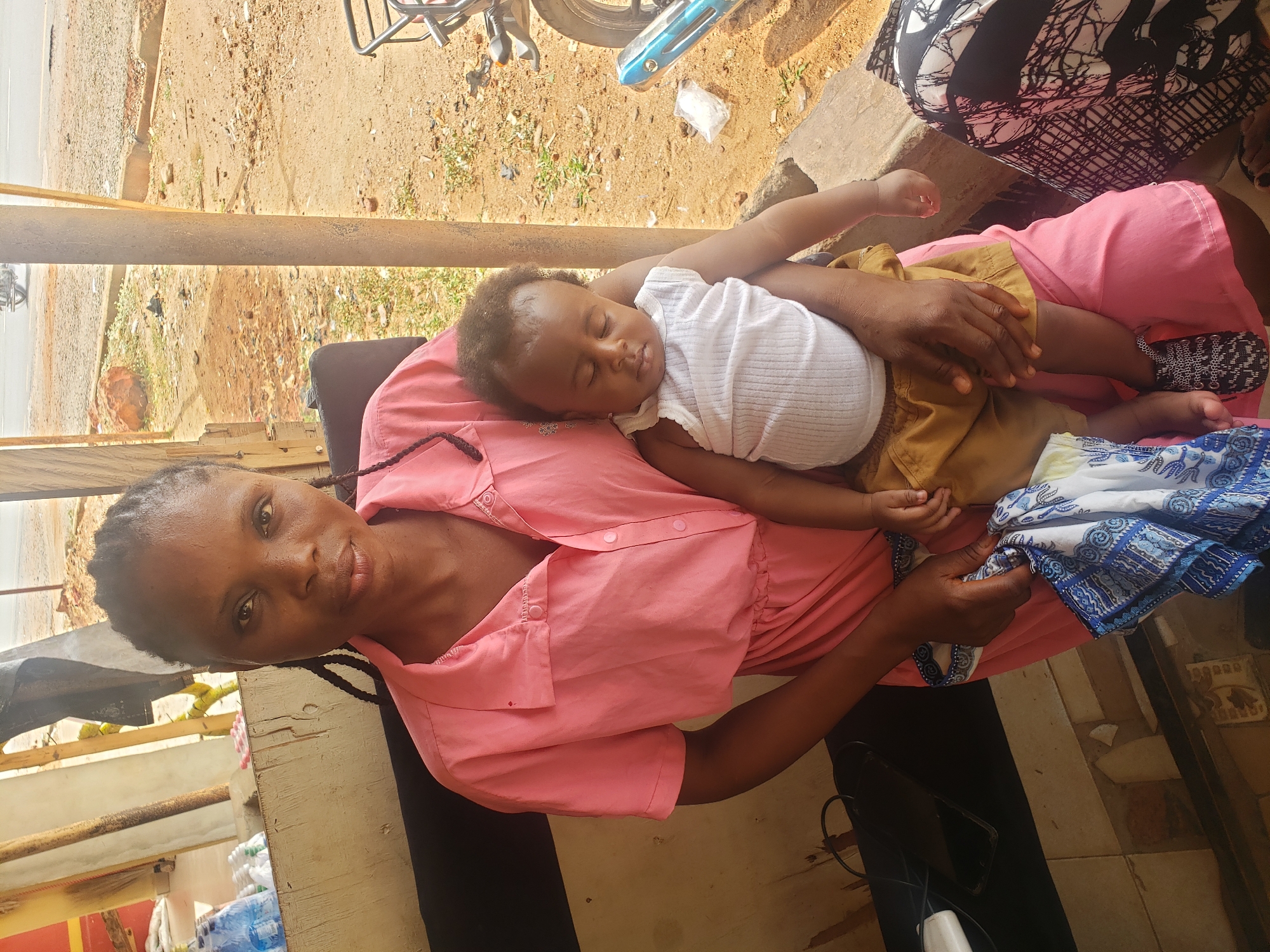 This portrait-style image, taken in Adankolo, Nigeria, features a woman holding a sleeping infant in an outdoor or semi-outdoor setting during bright daylight. The woman, with dark skin and braided hair, looks directly at the camera with a calm expression, wearing a pink, short-sleeved collared dress.
The infant, also with dark skin and curly dark hair, is sound asleep on the woman's lap and chest. The baby is dressed in a white ribbed sleeveless top and tan or khaki shorts. One foot is visible, wearing a black and white patterned fabric or shoe. The woman gently supports the baby, with one hand near the baby's upper back and the other holding a blue and white patterned cloth by the baby's leg.
The background suggests a roadside or market area. To the left, there's a dirt road with some sparse vegetation and parts of a metallic object, likely a motorcycle or bicycle. The ground transitions to a more paved or tiled surface closer to the subjects. Visible objects include stacked plastic bottles of water, a plastic bag with white contents, and wooden planks or structures. A person wearing black and white patterned clothing is partially visible in the upper right. The strong sunlight indicates a sunny day. No visible text is discernible in the image. The scene captures a peaceful moment of rest for the mother and child amidst their surroundings.