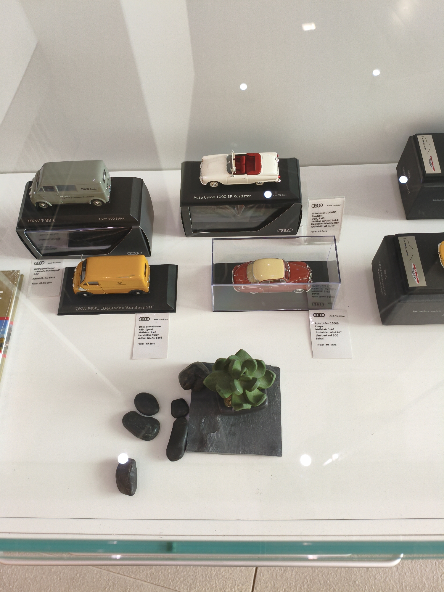 The image displays a collection of model cars in a glass display case. The models appear to be of vintage vehicles, including a green van with "DKW Kombi" text, a white convertible with a red interior, and a red and tan coupe. Each model is presented on a black base with a label providing details such as the model name, scale, manufacturer, article number, and price. The labels indicate that these are "Audi Tradition" collectibles, with some being limited editions. The display case is clean and well-lit, with reflections visible on the glass. A small succulent plant and dark stones are placed on a slate tile in the foreground of the display. The overall environment suggests a retail setting, possibly a gift shop or museum store in Ingolstadt, Germany, a city historically significant to Audi.