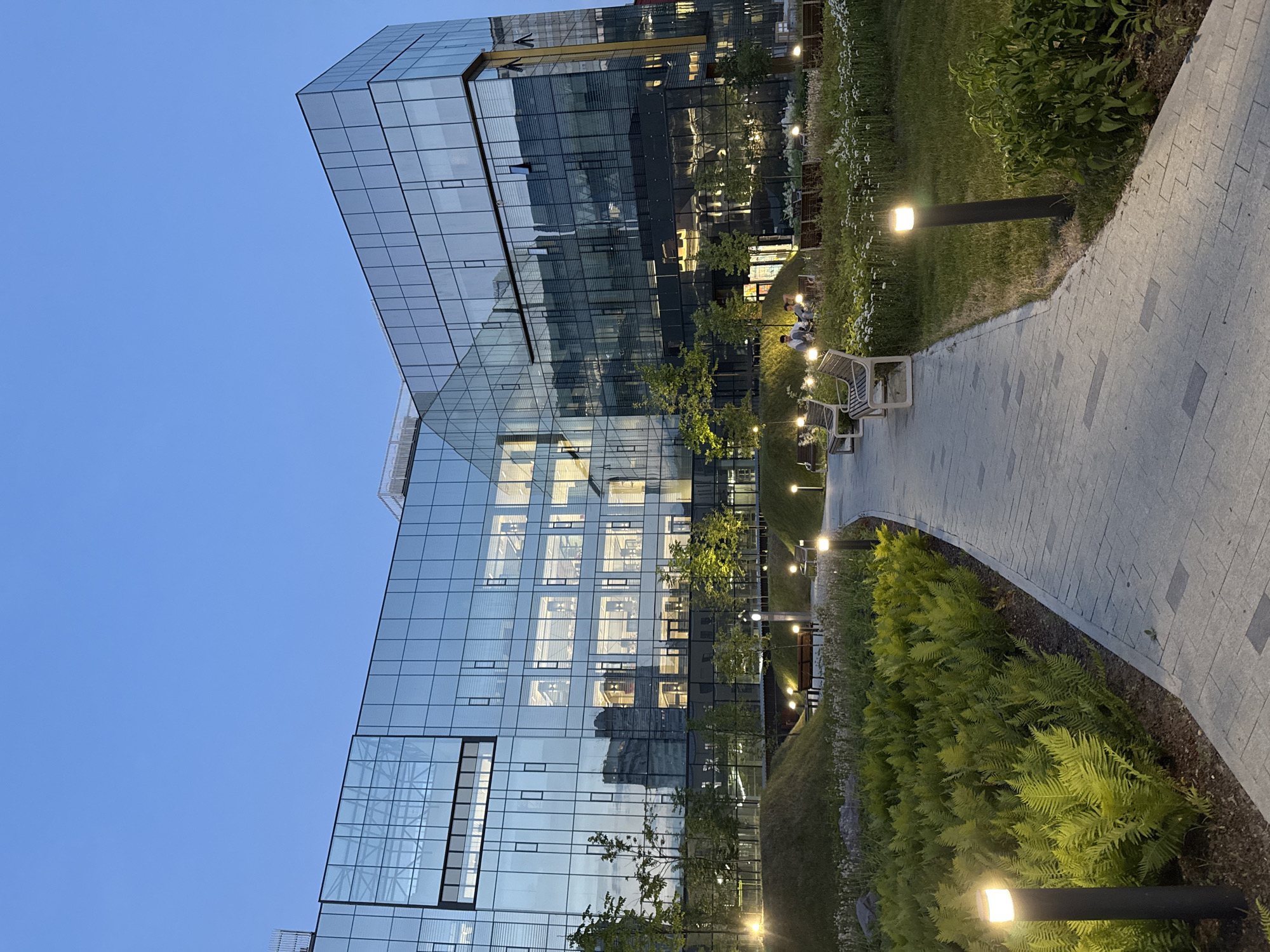 This image depicts the modern glass-facade building of Sberbank's "Dom 12" innovation center, located in Moscow, Russia. The scene is set during twilight, with the building illuminated from within, showcasing its multi-story interior through its reflective glass exterior. In front of the building, a landscaped park area features a paved walkway, benches, and lush greenery, including ferns and small trees. Several modern, low-lying lamps line the walkway, casting a warm glow on the path and surrounding vegetation. In the middle ground, two individuals are seated on a bench, seemingly engaged in conversation, enjoying the evening ambiance. The sky above is a clear, deep blue, transitioning from day to night.