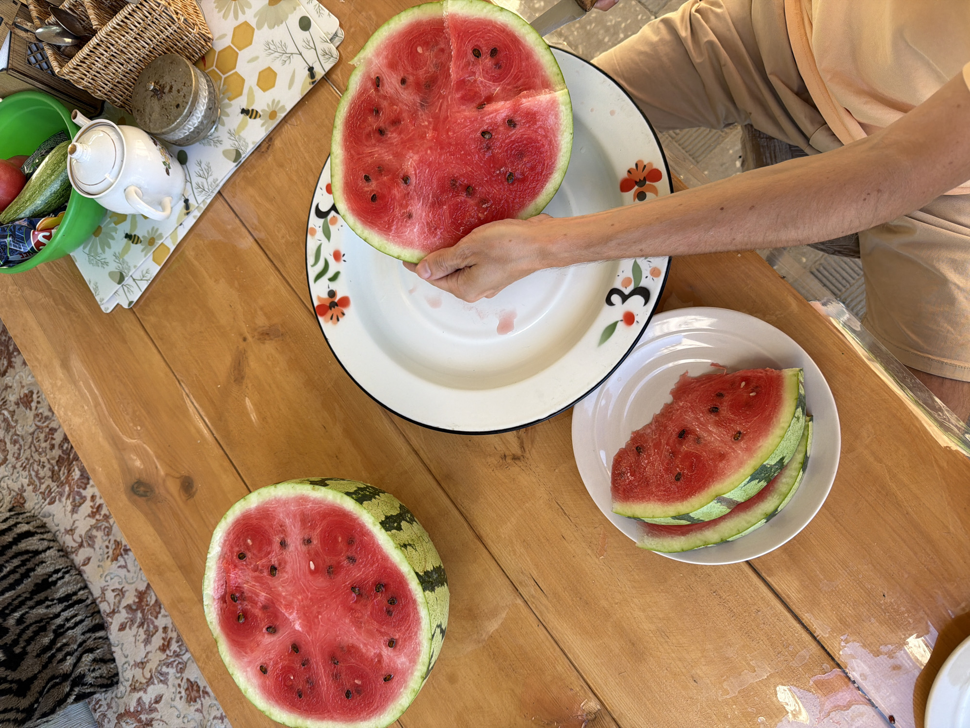 The image depicts an outdoor scene on a wooden table, possibly in Kamyshin, Russia, given the context. A person's arm and lap are visible in the lower right portion of the frame. The table holds several items related to preparing and consuming watermelon. On the left, a halved watermelon, its red flesh and black seeds exposed, rests on the table. To the right of the watermelon, a white plate with floral decoration holds a few slices of watermelon. A larger, decorative plate, also with floral accents, is positioned centrally, and a hand is holding a slice of watermelon over it, with a knife visible on the edge of the plate. In the background, other items are present, including a green bowl containing vegetables and a small white teapot. The overall impression is of a casual gathering or meal being prepared outdoors.