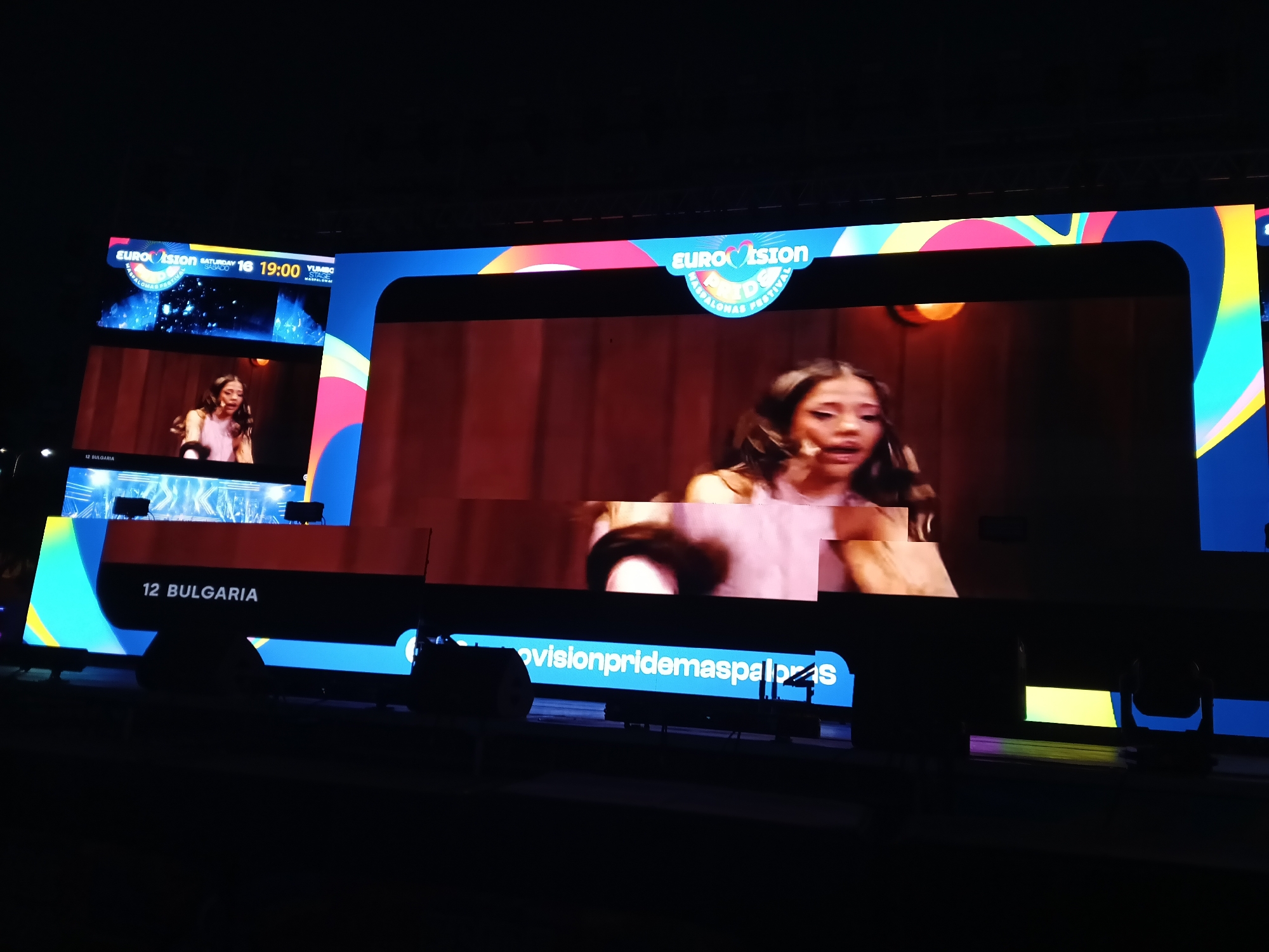 A large screen displays the Eurovision Maspalomas Pride Festival in San Bartolomé de Tirajana, Spain. A woman with long dark hair and a pink sleeveless top is singing on screen. The name "12 BULGARIA" is visible. The date and time "SATURDAY 16 19:00" are displayed on the screen. The text "ovisionpridemaspalomas" is partially visible on the bottom of the screen. Other lighting equipment is visible under the screen.