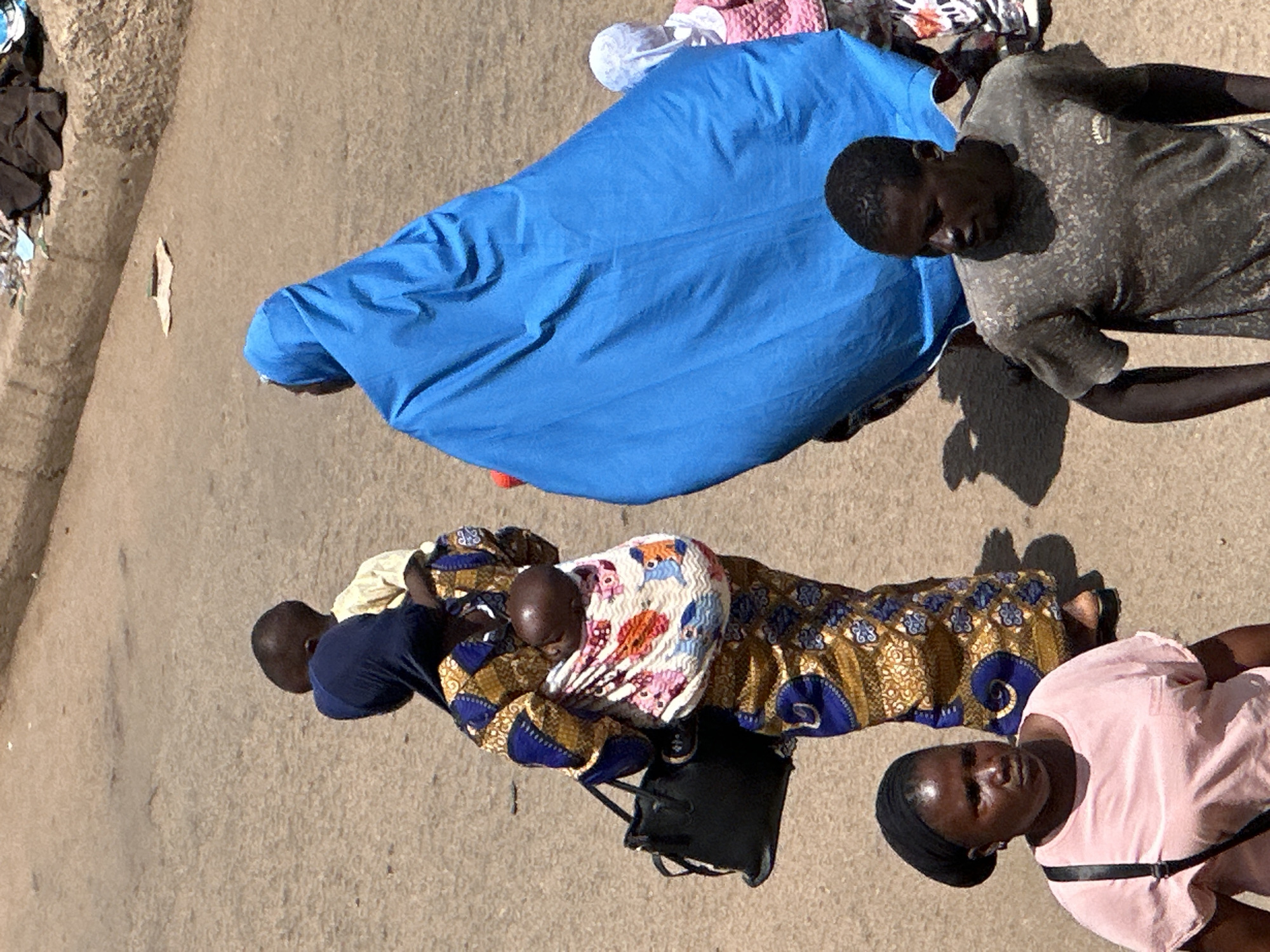 In Bukuru, Nigeria, a medium shot captures a street scene with several individuals. In the foreground left, a Black woman wearing a light pink t-shirt and a black headscarf is looking to her right. Behind her and to her right, a Black man in a speckled olive-green t-shirt is looking to his right. Dominating the center of the frame, a Black woman in a vibrant yellow and blue patterned traditional dress, with a dark headscarf, is carrying a baby on her back wrapped in a colorful fabric sling. The baby, facing away from the camera, has dark skin and hair. To the right of this woman, another Black person, mostly obscured by a large, bright blue garment, is walking away from the camera. Partially visible on the far right is another individual wearing a white head covering and a pink patterned top. The ground is paved with a sandy, textured surface, and in the upper right background, a raised concrete ledge is visible with some debris.
