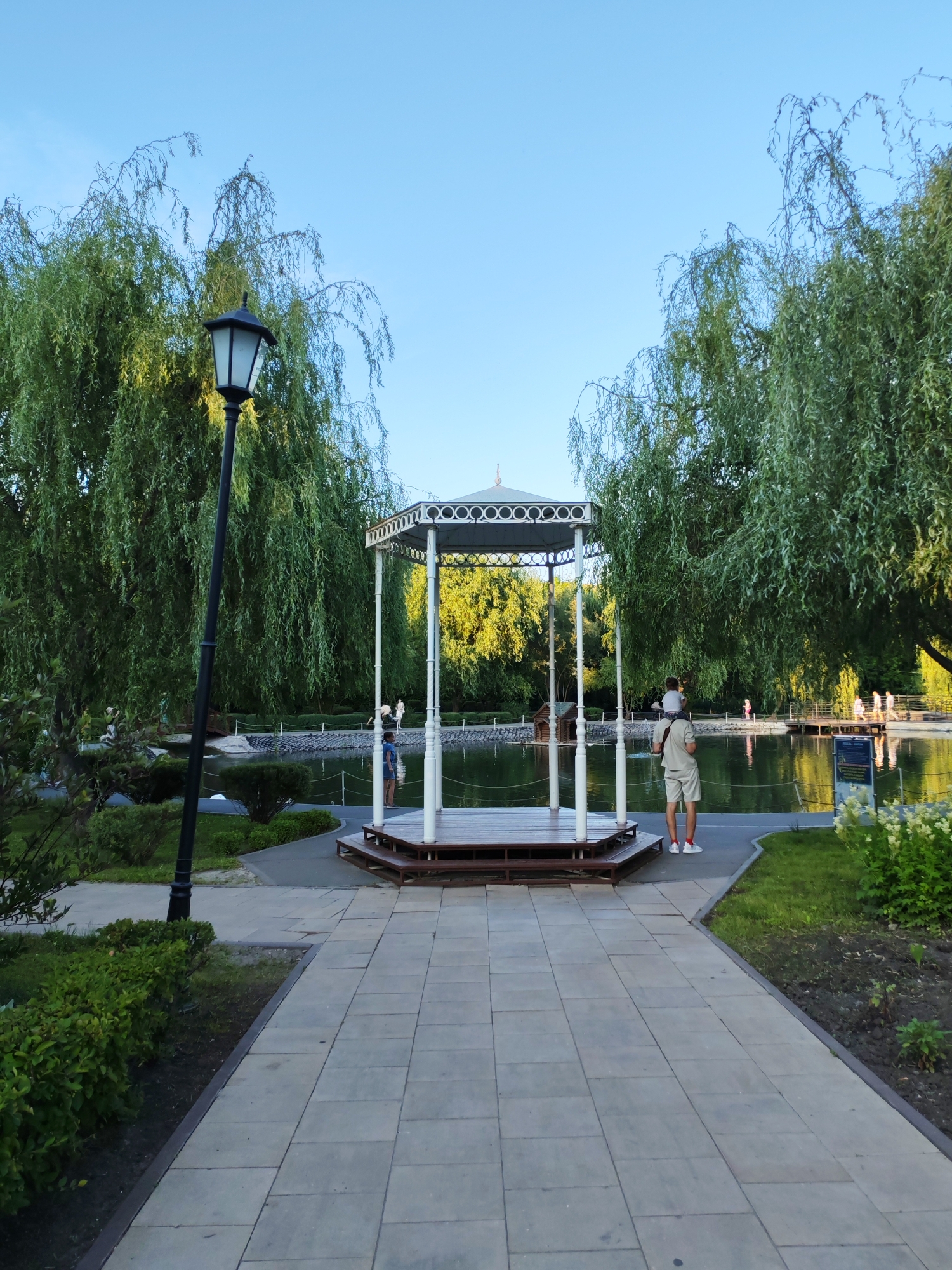 The image depicts a tranquil park scene in Volsk, Russia. A paved pathway leads towards a decorative gazebo situated next to a serene pond. The gazebo features a white, ornate roof supported by white columns, with a wooden platform. The pond is surrounded by a stone embankment and lush green trees, predominantly willows with their characteristic drooping branches, which cast gentle shadows on the water.
In the foreground, a black lamppost stands to the left of the pathway. Two individuals are visible near the gazebo. On the right, a person wearing light-colored shorts and a t-shirt stands with their back to the camera, holding a child on their shoulders. The child is wearing a white t-shirt. To the left of the gazebo, closer to the pond, a person in blue shorts is standing. In the background, several other figures can be seen walking along a path on the opposite side of the pond, suggesting a public park setting. The sky is clear and blue, indicating fair weather. The overall atmosphere is peaceful and idyllic, characteristic of a summer day in a city park.