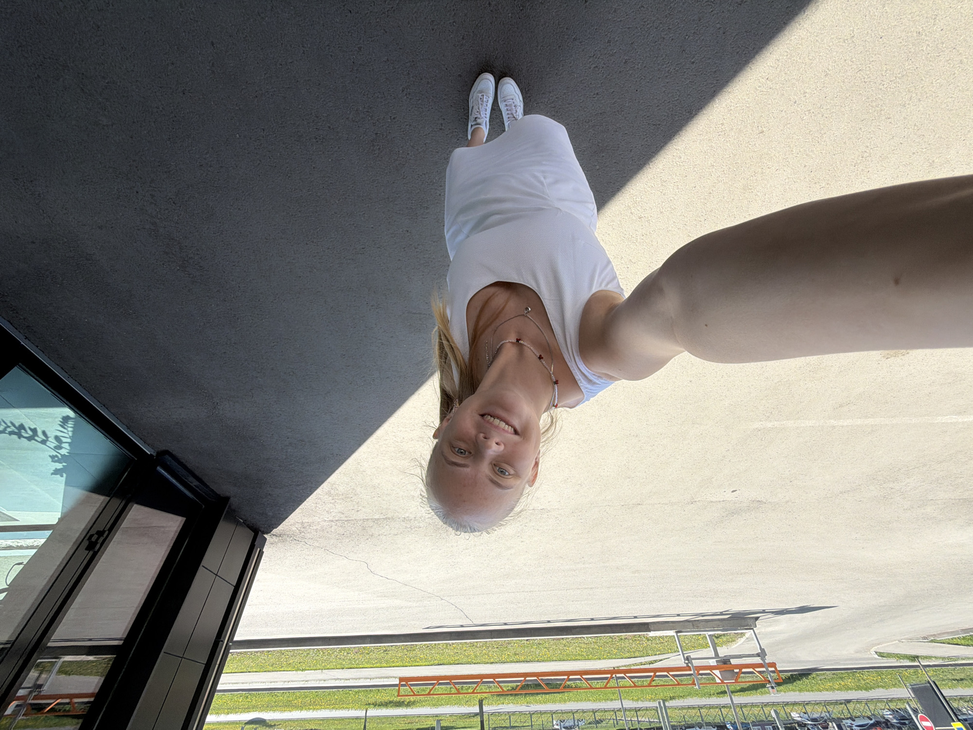 A person, presumably female, in a white dress and white sneakers is taking a selfie. She is standing on a light-colored concrete surface with a dark overhanging structure to her left. A strip of grass and parked cars are visible in the background. The location might be Izhevsk, Russia, based on the user prompt, but there are no specific visual indicators in the image to confirm this location.