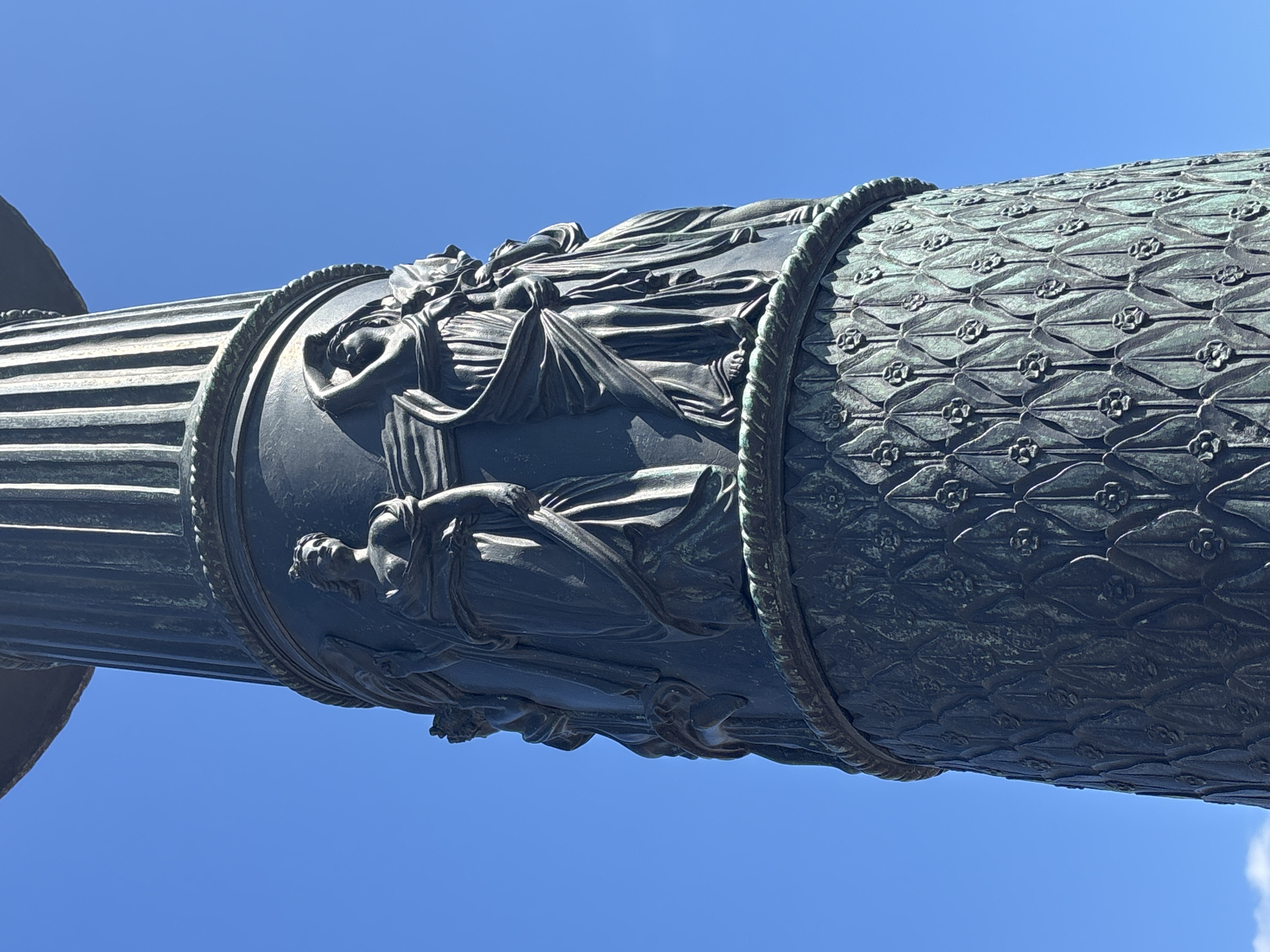 The image shows a close-up of a dark bronze column with intricate relief sculptures depicting classical figures. The column is part of a monument in Saint Petersburg, Russia. The figures are draped in flowing robes, and some appear to be holding or interacting with each other. The lower section of the column is decorated with a repeating pattern of stylized leaves and floral motifs. The sky above is a clear, bright blue. This column is part of the monument to Catherine the Great in Nevsky Prospekt, Saint Petersburg, Russia.