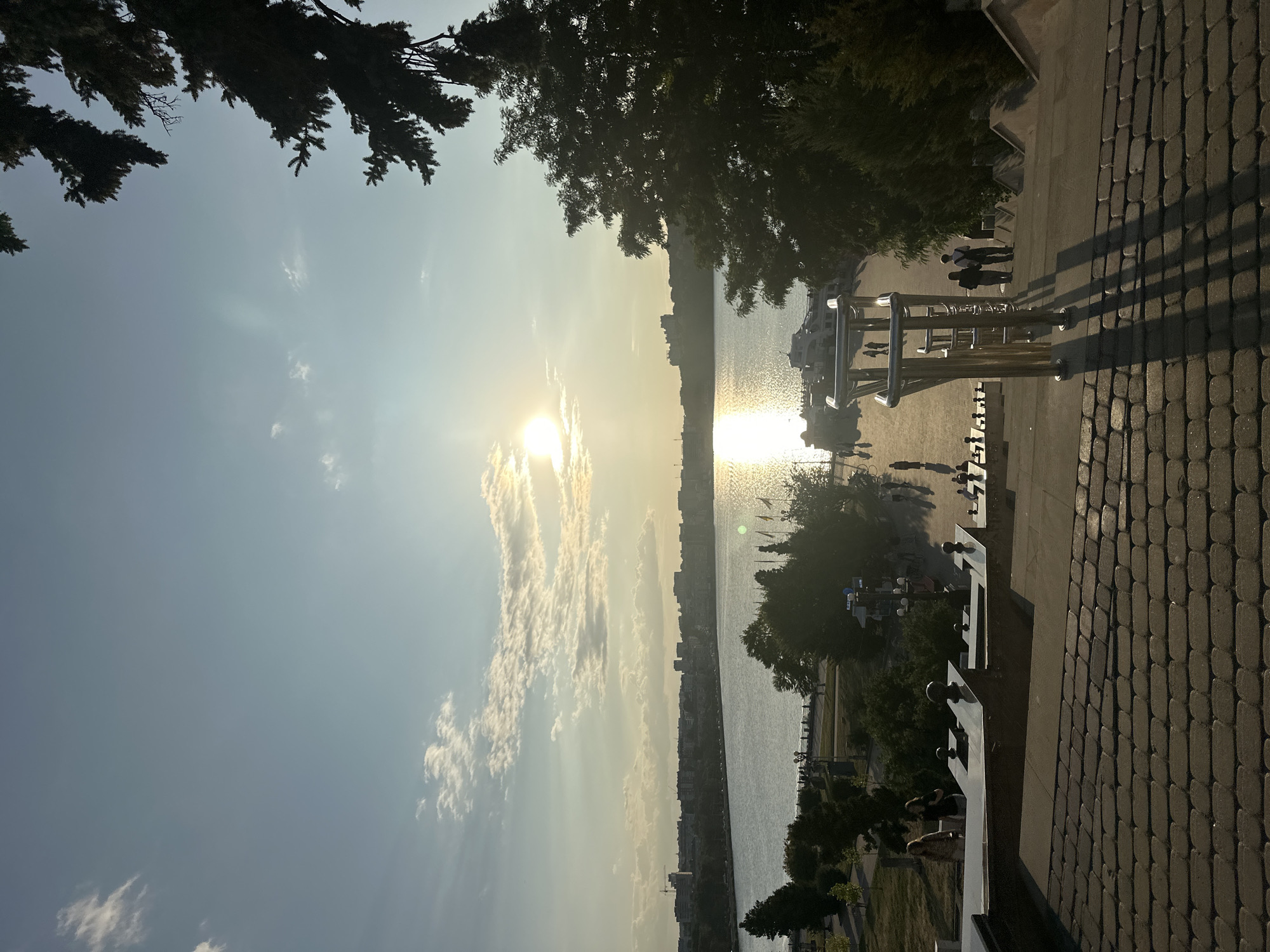 The image depicts a view of the Ternopil pond in Ternopil, Ukraine, at sunset. In the foreground, a wide staircase made of dark paving stones leads down towards the water. A metal railing with a modern design runs along the right side of the stairs, casting long shadows on the steps. Several people are visible walking down the stairs, their silhouettes dark against the light.
Beyond the stairs, a paved walkway stretches along the edge of the pond. Several trees, including a large evergreen on the right, line the walkway and the pond's edge. The pond itself reflects the warm glow of the setting sun, creating a shimmering path of light across its surface. In the distance, the cityscape of Ternopil is visible, with buildings lining the far bank of the pond. A white boat is docked along the waterfront. The sky is a pale blue with scattered white clouds, illuminated by the bright, low-hanging sun. The overall atmosphere is serene and picturesque.