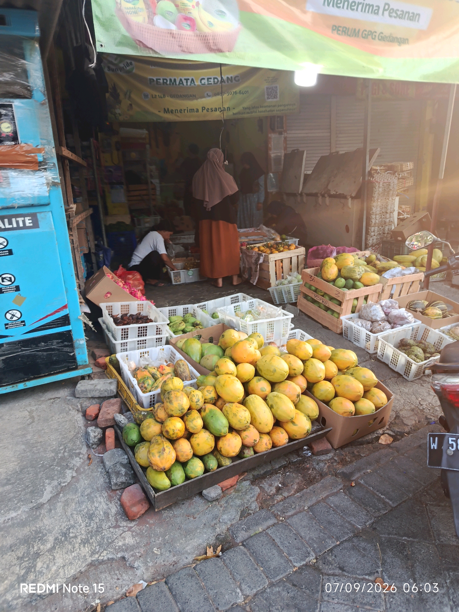 The image shows an outdoor fruit stand in Keboansikep, Indonesia, likely in front of a shop. Papayas are prominently displayed in baskets and wooden crates in the foreground and middle ground. Other fruits, possibly rambutan or lychee, are in white plastic crates. A person wearing a hijab is partially visible in the background, standing near the shop entrance. Another person is crouched down inside the shop. The shop has a banner above its entrance that reads "PERMATA GEDANGAN" and mentions "Menerima Pesanan," indicating it accepts orders. The ground is paved with concrete and bricks. A portion of a motorcycle is visible on the right side of the image, with a license plate starting with "W 58". The date stamp "07/09/2026 06:03" and "REDMI Note 15" are visible in the lower corners of the image. The overall scene depicts a typical Indonesian fruit market.