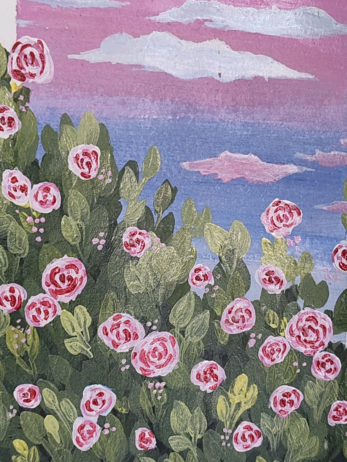 The image is a close-up, top-down view of a painting depicting a dense cluster of pink and white roses amidst lush green foliage. The background features a sky transitioning from a soft pink near the horizon to a vibrant blue, with scattered white and pinkish clouds. The painting's textured brushstrokes are visible, suggesting a vibrant, impressionistic style. There are no people, actions, or interactions depicted. The scene is entirely naturalistic and does not contain any elements that would indicate a specific location within the city of Mysuru, India.