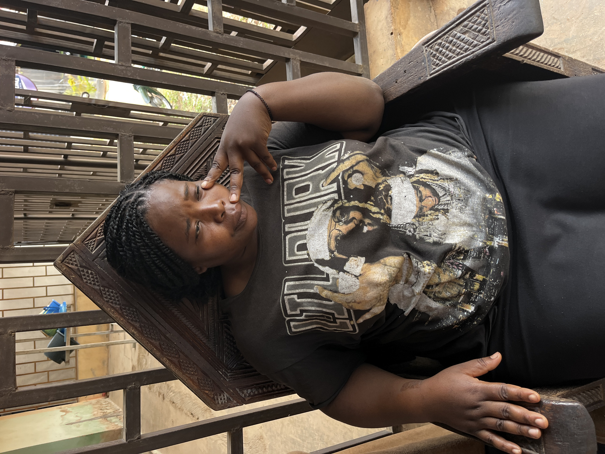 A medium shot captures a Black woman with dark braided hair sitting in a carved wooden chair. She is wearing a dark t-shirt with a graphic print of a man's face and text, and a black skirt. Her right hand is placed on her cheek with her fingers touching her temple, and her eyes are cast downwards. The chair is ornately carved with geometric patterns. Behind her, a metal railing structure with vertical bars is visible, and beyond that, a glimpse of an outdoor environment with some greenery and possibly a motorcycle. The scene appears to be an outdoor or semi-outdoor covered area, possibly a porch or courtyard.