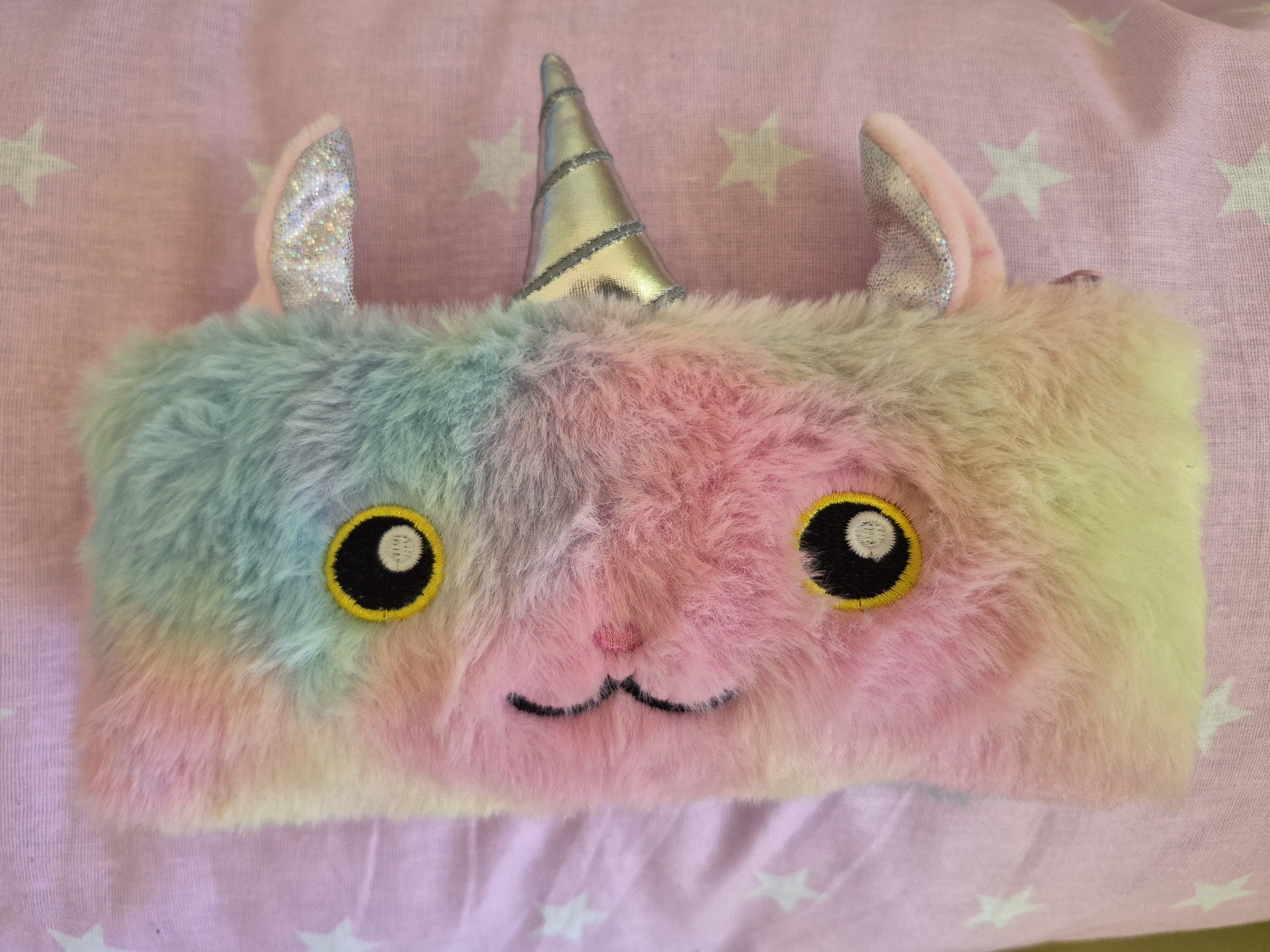 A close-up photograph shows a rectangular, plush, rainbow-colored unicorn pencil case lying on a pink fabric surface patterned with white stars. The pencil case features a silver unicorn horn, two silver ears, two round eyes with yellow stitching, and a small embroidered mouth. The photograph is taken indoors under diffused light. The environment shows the pencil case lying flat on the fabric. There are no people present in the image. There is no indication of this image being from Tobolsk, Russia.