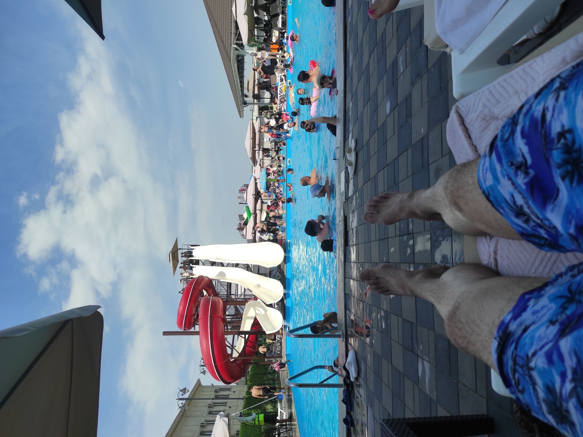 The image depicts a lively outdoor water park scene on a sunny day. In the foreground, two pairs of legs are visible, one wearing blue patterned swim trunks and resting on a lounge chair. Behind these legs, a bright blue swimming pool is filled with numerous people of various ages enjoying the water. Children and adults are swimming, some with inflatable rings, and several are clustered around the edge of the pool.
To the left of the pool, a multi-level water slide structure with red and white slides dominates the midground. People are visible at the top of the slides, preparing to descend, and others are in the pool at the bottom of the slides. A building with light-colored walls and windows is situated to the far left, with some trees and greenery. Numerous umbrellas provide shade around the pool area.
The sky is a clear blue with scattered white clouds. The ground around the pool is paved with dark and light square tiles. The overall atmosphere is one of a recreational resort or a public swimming facility. There is no visible indication of the specific location being in the city of Шалқар, Kazakhstan.