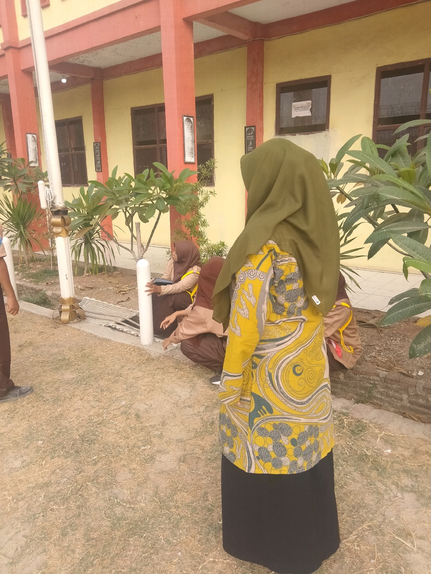 The image depicts an outdoor scene in front of a building with a yellow facade and red trim. A flagpole stands on the left. Several individuals, likely students in uniform, are present. One individual in the foreground wears a green hijab and a yellow top with a black skirt, displaying a paisley pattern. Two other individuals are seated on the ground in the middle ground, also appearing to be in uniform. The ground is covered with dry grass and dirt. In the background, the building features windows and signs, one of which reads "PERPUSTAKAAN" (Library) on a piece of paper taped to a window. The overall environment suggests a school or institutional setting in a tropical climate, possibly in Suka Sari, Indonesia, given the architectural style and vegetation. The lighting indicates it is daytime, likely sunny.