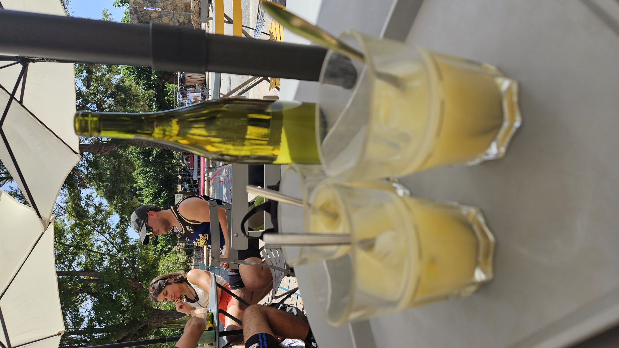 In Sant'Antonino, France, a group of people are seated at an outdoor cafe table under a large white umbrella. A man wearing a baseball cap and sunglasses is looking to his left. A woman with sunglasses perched on her head is looking down. Three glasses of yellow liquid, possibly a dessert or drink, are on a gray tray in the foreground, each containing a spoon. A green bottle of wine stands behind the glasses. The background shows trees and buildings, indicating a sunny day in a town or village setting.