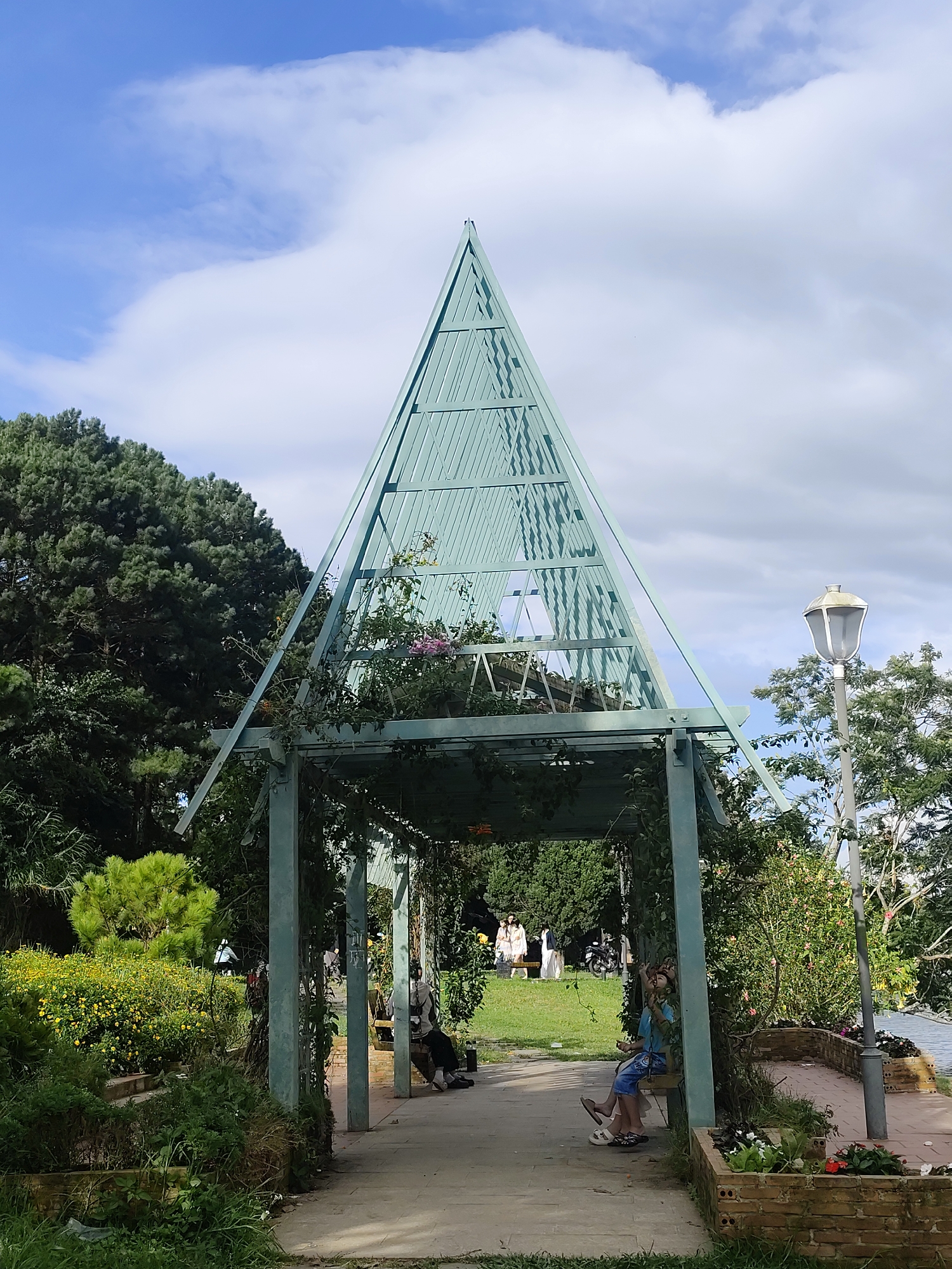 The image shows a triangular-shaped teal-colored structure with a metal frame, possibly a pavilion or trellis, in a garden setting. The structure is adorned with climbing plants, some of which have pink flowers. In the foreground, there is a paved walkway leading under the structure, with several people sitting on benches within the shaded area. One person, a child, is on a swing, looking upwards. Behind the structure, a patch of vibrant yellow flowers is visible, along with lush green trees. A lamppost stands to the right of the structure. In the background, a few people are walking on a grassy area, and a motorcycle is parked. The sky above is partly cloudy with a bright blue hue. This scene is likely located in the city of Da Lat, Vietnam, known for its gardens, parks, and French colonial architecture, which often features structures like this.