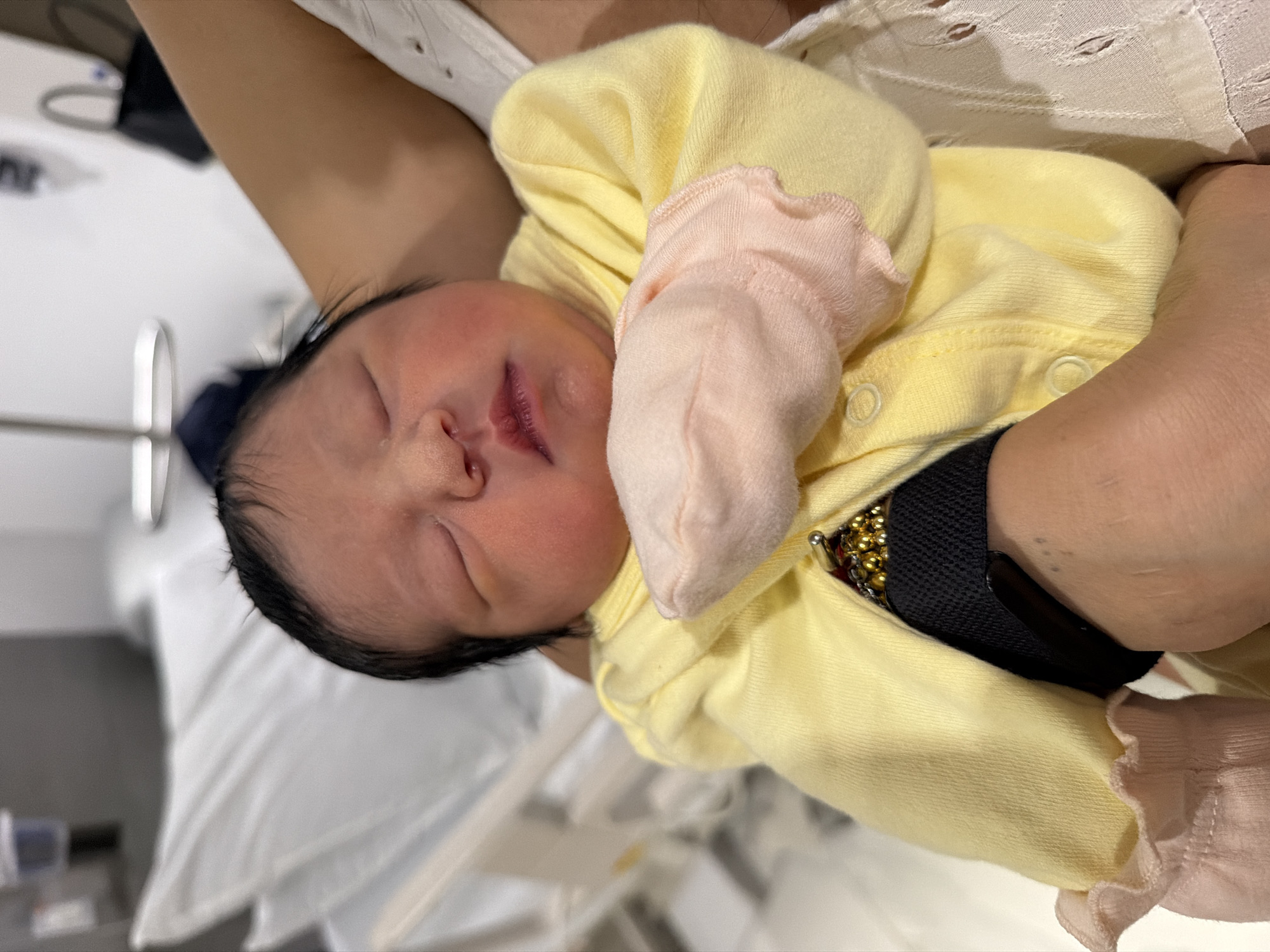 A newborn infant is being held by an adult. The infant is fair-skinned with black hair and is wearing a yellow outfit with pink mittens. The adult is wearing a white lace top. The background includes a hospital bed and medical equipment. It is not possible to ascertain the location as Villahermosa, Mexico, from the image provided.