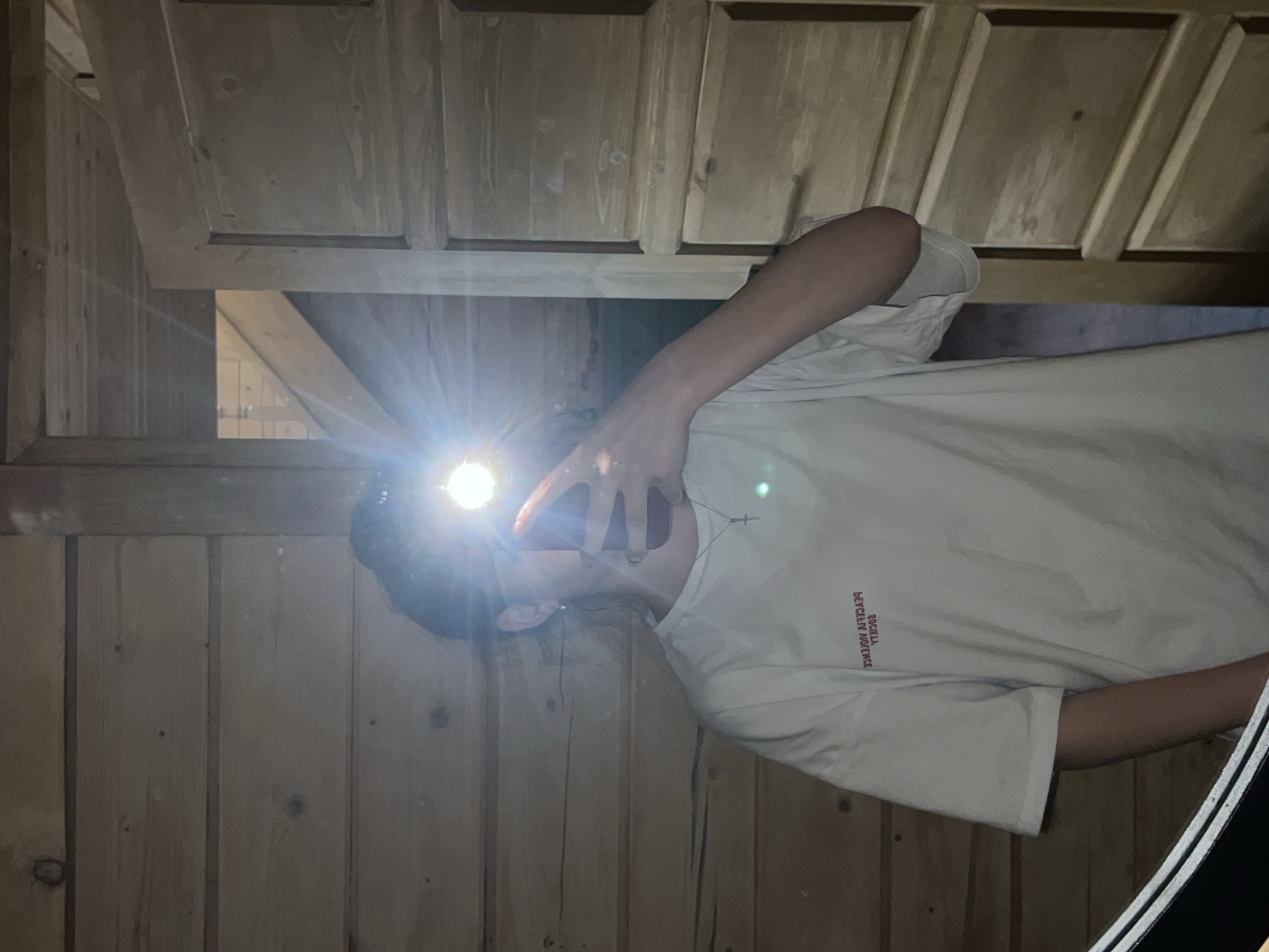 A person is captured in a mirror selfie within a room with wooden paneling and a wooden door. The individual is wearing a white t-shirt with a red graphic and a cross necklace. The flash from the camera is causing significant glare in the image, obscuring the person's face. The room appears to be dimly lit.