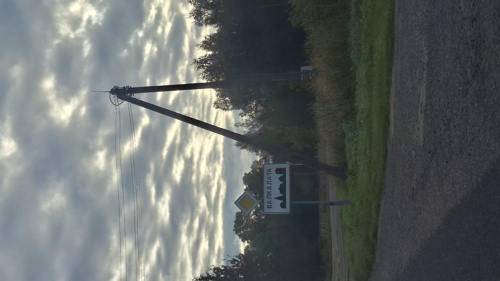 The image captures a street scene with a signpost in the foreground. The sign, written in Cyrillic script, reads "ВАЛКАЛАТА" and is accompanied by a silhouette of buildings. Above this sign, a yellow diamond-shaped sign is visible. The signpost is leaning against a tall, wooden utility pole that supports power lines. The background is dominated by a dense forest with the sky filled with dramatic, overcast clouds. A paved road runs through the foreground, curving to the left. The setting appears to be rural, possibly a village entrance given the town name sign. The lighting suggests it is daytime, possibly early morning or late afternoon, due to the diffused light filtering through the clouds.