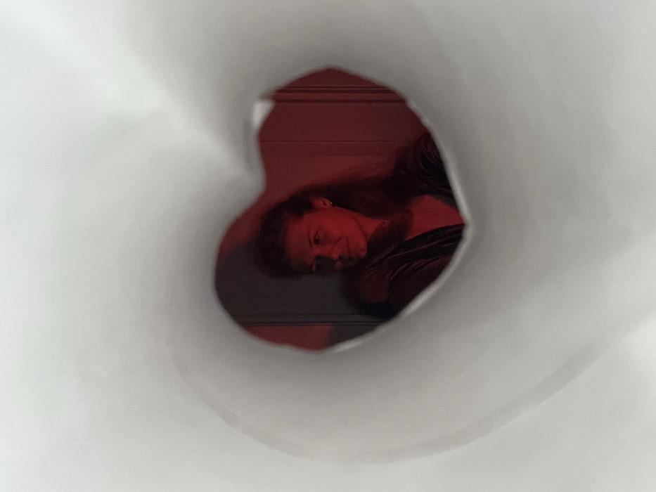 A woman with long brown hair and light skin peers through a heart-shaped opening. She's wearing a dark top, and her face is illuminated with a red light. The background behind her is also red. The heart shape is framed by a white, smooth material, creating a tunnel-like effect, focusing the viewer's attention on the woman's face. The image has a dreamlike or surreal quality due to the lighting and perspective.