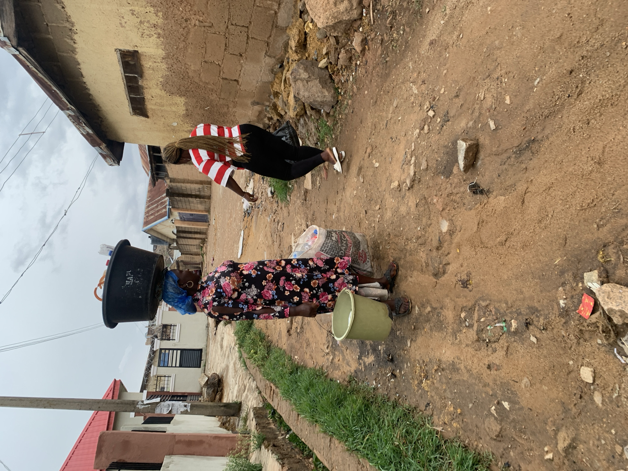 The scene is an unpaved street in Jos, Nigeria. An African woman with a blue headscarf and floral dress carries a large black bucket on her head and a light green bucket in her right hand. A large bag or package is next to her. Another woman, wearing a red and white striped shirt and black pants, stands nearby. Buildings flank the street, with some appearing unfinished or in disrepair. The ground is dirt and scattered with debris.