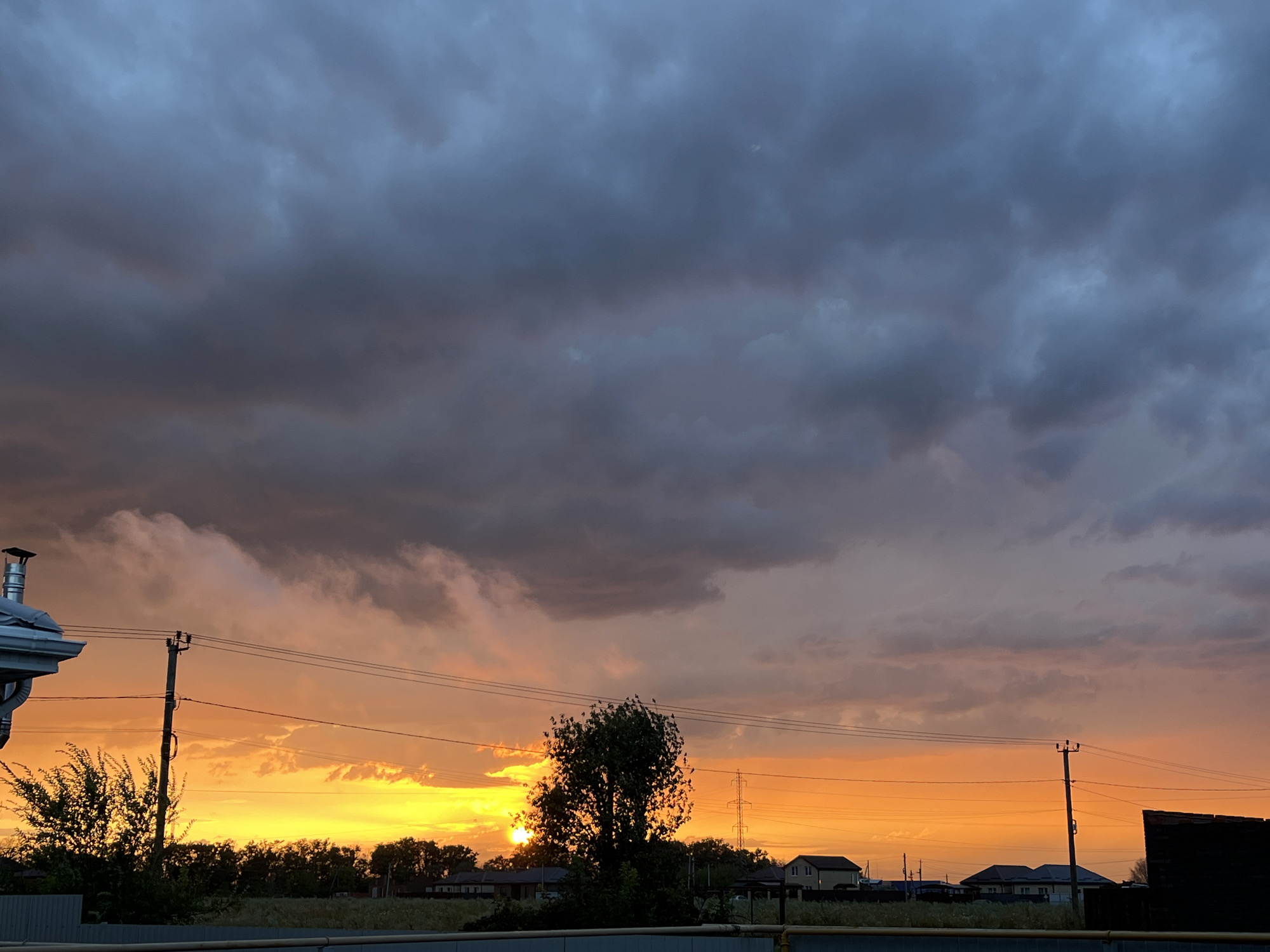 The image captures a sunset over a rural or suburban area, likely in the Taganrog City District of Russia. The sky is dominated by dramatic clouds, with a band of vibrant orange and yellow stretching across the horizon, indicating the setting sun. Above this bright band, the clouds transition to darker shades of gray and blue, suggesting an approaching storm or the deepening twilight.
In the foreground, on the left side of the image, a section of a building with a silver gutter and a chimney is visible. To the right of the building, a single utility pole with wires strung between it and another pole further down the line is present. A tree with sparse foliage stands in the mid-ground, partially silhouetted against the bright sky. A line of trees forms a dense border behind the foreground elements, and beyond that, several houses with pitched roofs are scattered across the landscape. A tall lattice tower is visible in the far distance. A metal fence runs along the bottom of the image, separating the viewer's vantage point from the scene beyond. There are no people visible in the image. The overall atmosphere is one of dramatic natural beauty, with the warm hues of the sunset contrasting with the ominous storm clouds.
