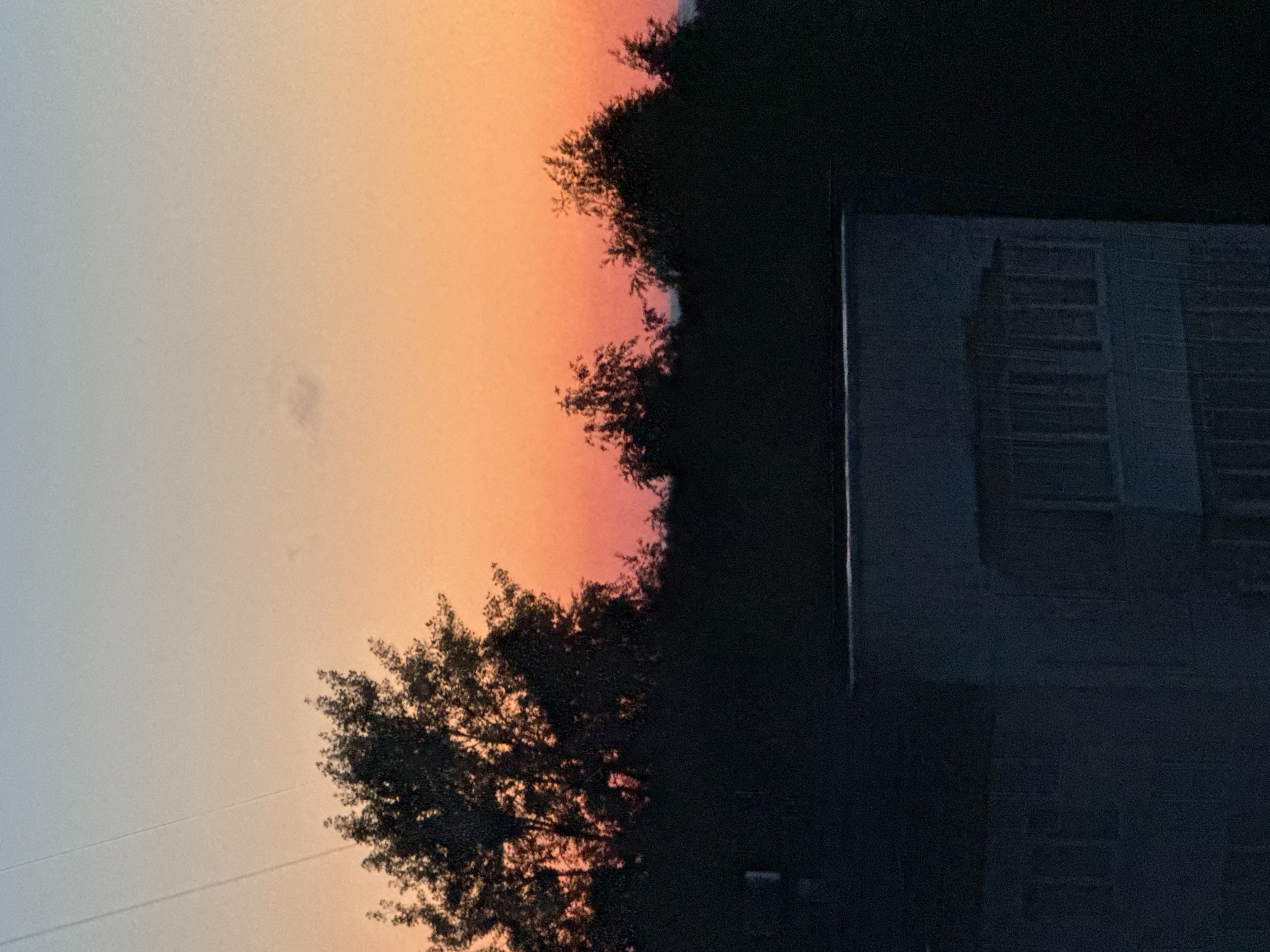 A photograph taken from a high vantage point shows a panoramic view of the city of Chaikovsky, Russia, during sunset. The sky is a gradient of colors, transitioning from a pale blue at the top to a vibrant orange and pink hue near the horizon. Silhouetted against the colorful sky are the dark shapes of trees and the top portion of a building with multiple windows. A faint contrail from an aircraft is visible in the upper left corner of the sky. The overall scene depicts a serene evening in an urban landscape.