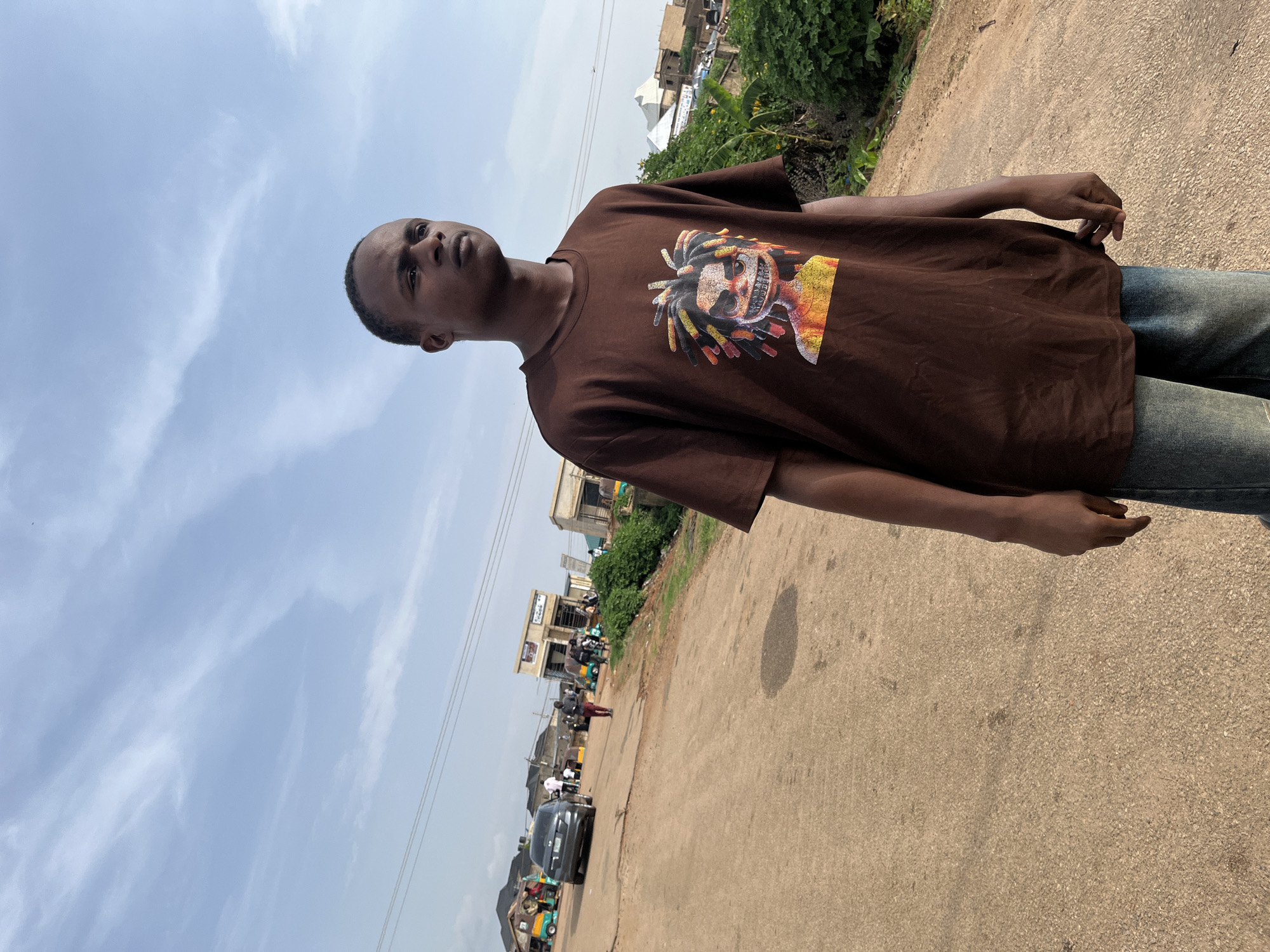 The image depicts a young man standing on a dirt road in Rafin Par, Nigeria. He is wearing a brown t-shirt with a graphic print and blue jeans. The man is looking to his right, with a neutral expression. The background shows a street scene with several buildings, including shops and houses. There are also vehicles such as cars and tricycles present, along with a few other people walking in the distance. The sky is partly cloudy. Lush green vegetation is visible on the right side of the road, bordering the scene. The road surface is primarily dirt with some patches of asphalt.