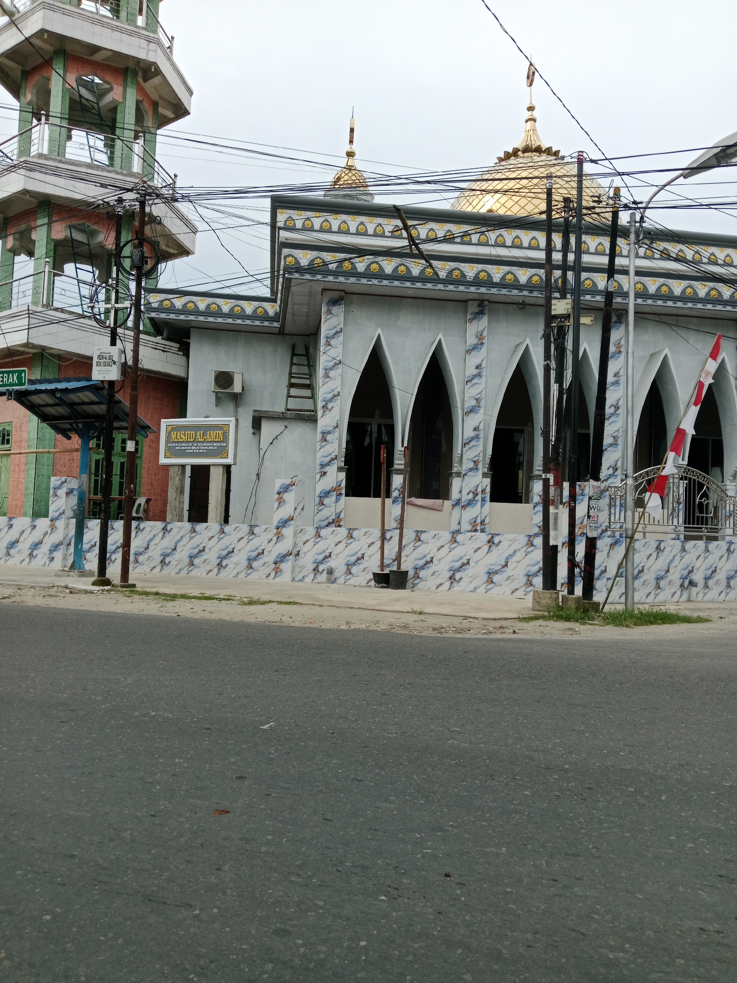 The image depicts the Masjid Al-Amin in the City of Binjai, Indonesia, viewed from across a paved road. The mosque features a light blue facade with arched windows and decorative tiling. A golden dome crowns the structure, topped with a crescent moon finial. To the left, a multi-tiered minaret with green and pink accents is visible. Overhead, a dense network of electrical wires crisscrosses the sky, supported by utility poles. A sign in front of the mosque reads "MASJID AL-AMIN". A red and white Indonesian flag is attached to a metal fence. The foreground shows the asphalt road and a small patch of grass. The sky is overcast.