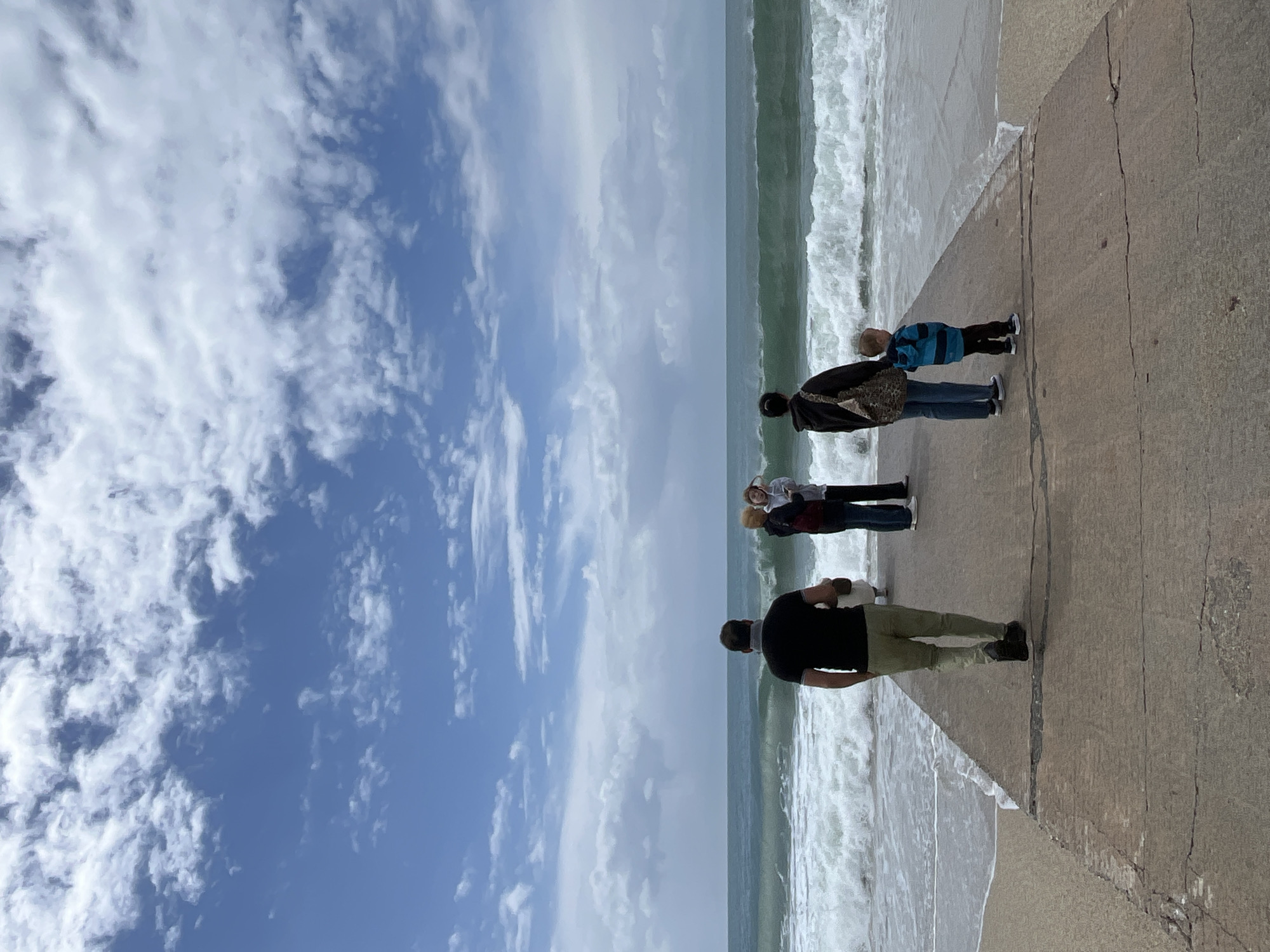 The image captures a group of four people standing on a concrete pier or edge overlooking the sea. The sea is choppy with waves, and the sky is blue with scattered white clouds. The people appear to be observing the waves. The location is the city of Alanya, Turkey.
A man is standing closest to the viewer, dressed in a black shirt and khaki pants. Behind him, a group of two adults and a child are standing. One adult has a black jacket on. The child has a blue and black jacket. The group is standing near the edge of the pier, with the sea waves visible in the background. The concrete pier has a prominent crack.