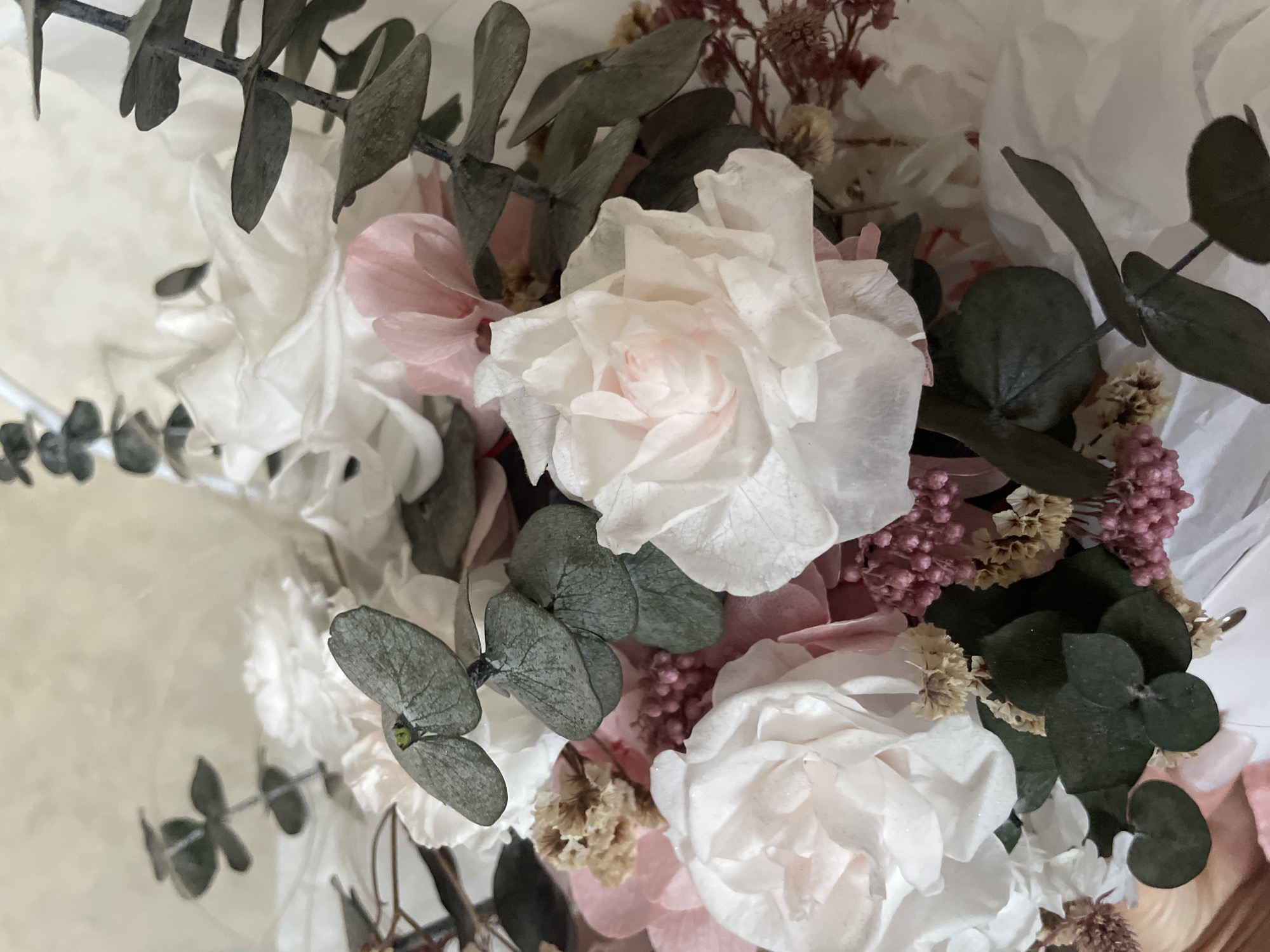 The image is a close-up, eye-level shot of a bouquet of dried flowers. The bouquet features several white and pale pink roses, interspersed with small clusters of pink and brown dried flowers. Dark green eucalyptus leaves are also present, adding a textural contrast. The arrangement appears to be set against a soft, out-of-focus background of white paper or fabric. There are no people or discernable landmarks that would indicate a specific location in Minsk, Belarus. The focus is entirely on the floral arrangement itself.