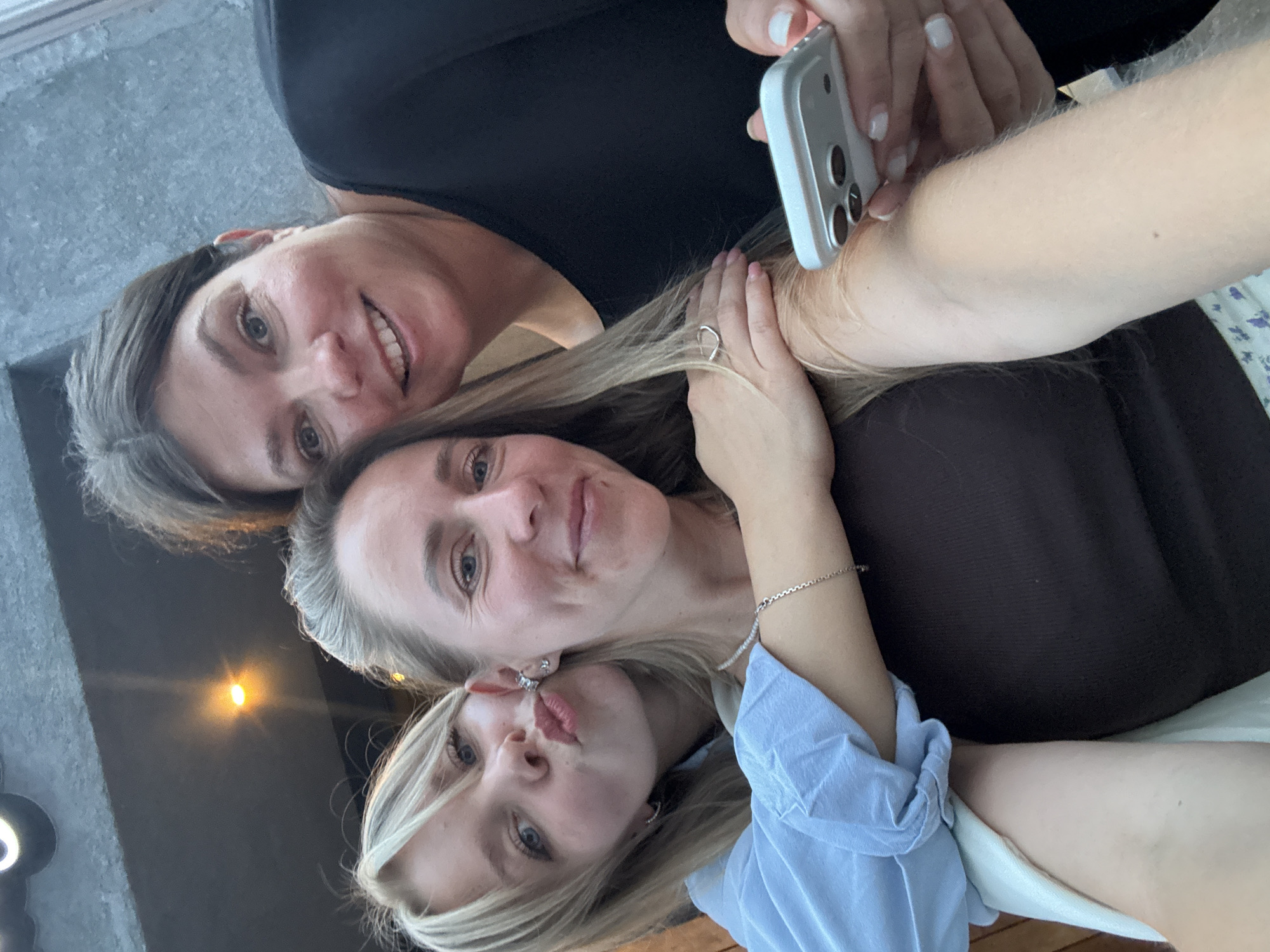 Three women are posing for a selfie outdoors. The woman on the left, who appears to be a child, is leaning forward with her lips puckered. The woman in the middle is smiling and has her arm around the child. The woman on the right, who is holding a smartphone, is also smiling. The background is blurry but appears to be an outdoor setting with a wall and some lighting. The image does not contain any information or visual cues that would allow for a specific location within the city of Chaikovsky, Russia, to be identified.