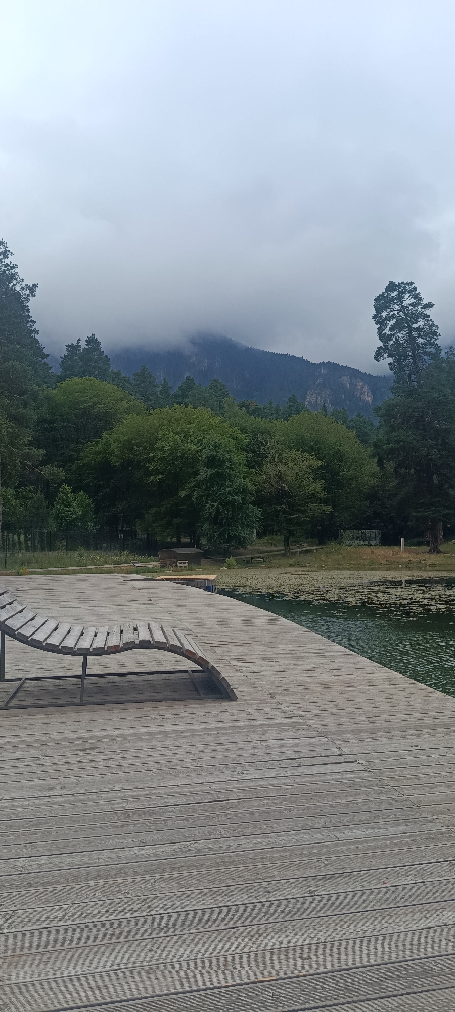 A wide shot captures a wooden deck that curves into the frame from the left foreground, leading towards a body of water. A single, empty, weathered wooden lounge chair rests on the deck, angled to face the water. The water, visible in the mid-right of the image, is dark green and covered in scattered water lilies. Beyond the water, a dense forest of green trees creates a thick backdrop. In the distance, mountains rise, partially obscured by heavy, gray clouds that blanket the sky. The scene is set in Teberda, Russia, a location known for its mountainous landscape and natural beauty.