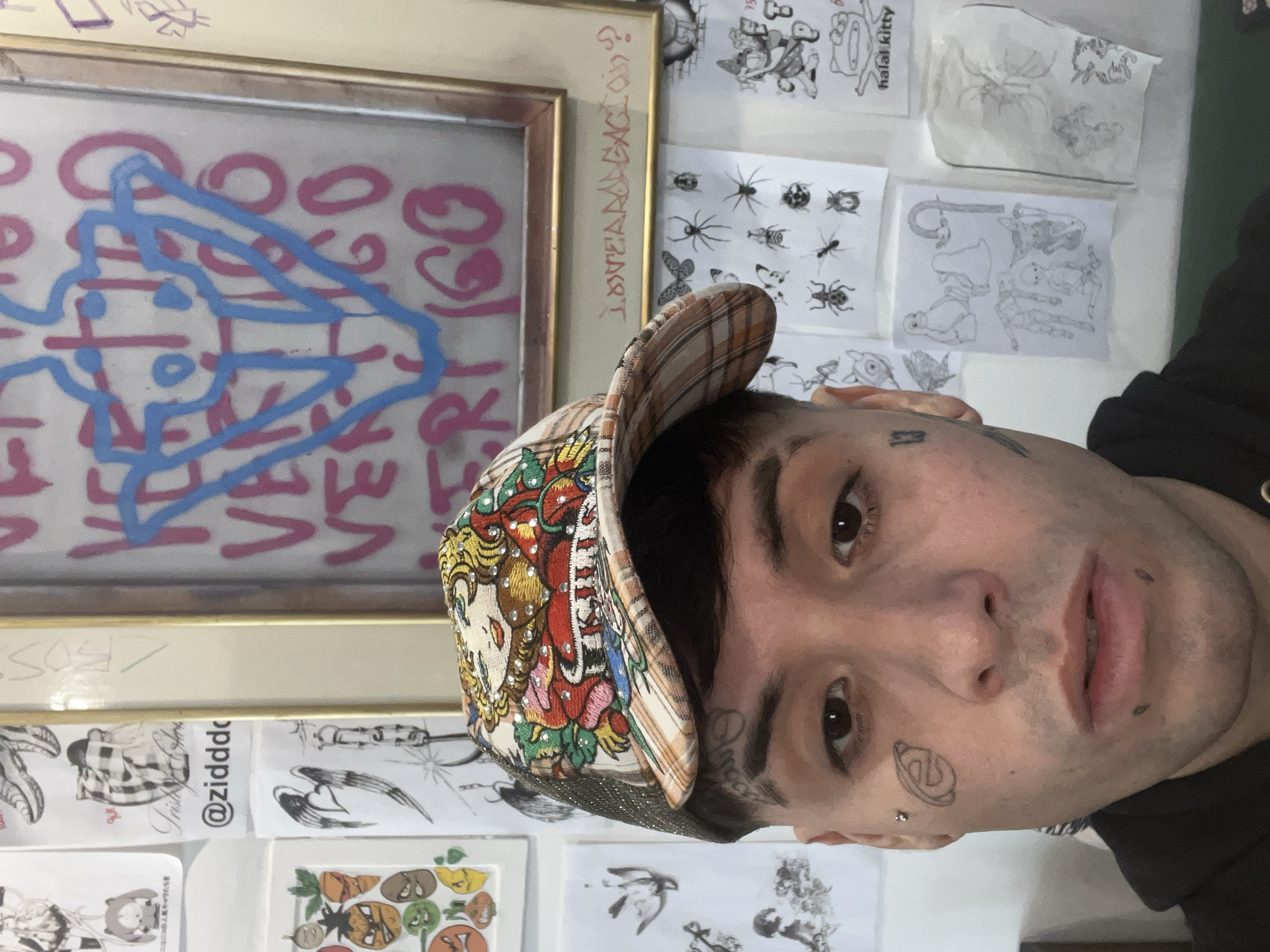 A person with tattoos on their face is wearing a patterned cap. In the background, there's a framed drawing with blue and pink markings, along with several other drawings and sketches pinned to a wall. One of the pinned drawings has the text "halal kitty". Another says @zidddd. There are some text markings on the frame which say things like "VERTIGO" and "FRT 160" and "BOS" and "VARTIS". The context or specific location within Buenos Aires is not discernible from this image alone.