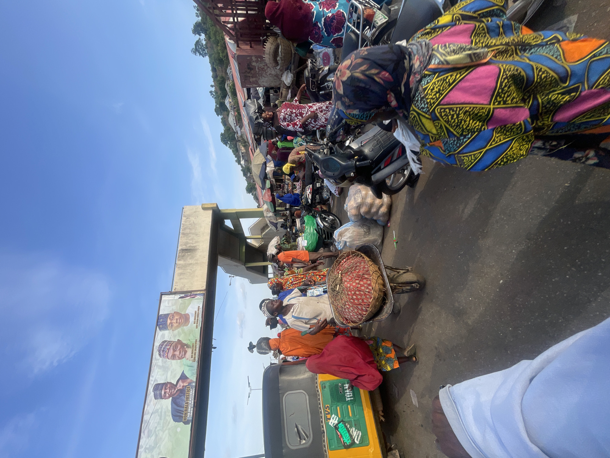 This image depicts a bustling scene in Lokoja, Nigeria. The area seems to be a market or a busy street lined with vendors. There are many people present, some wearing colorful African prints. Several motorcycles are parked in a row, some loaded with goods or belongings. There is a three-wheeled vehicle (Tuk-tuk) visible with its distinctive yellow and green coloring. Some vendors are carrying goods on their heads. An overhead sign displays political campaign posters for HON BALA JUSTICE, HIS EXCELLENCY ELDER JOEL SALIFU OYTHO, and HIS EXCELLENCY LOV USMAN AHMED UND.