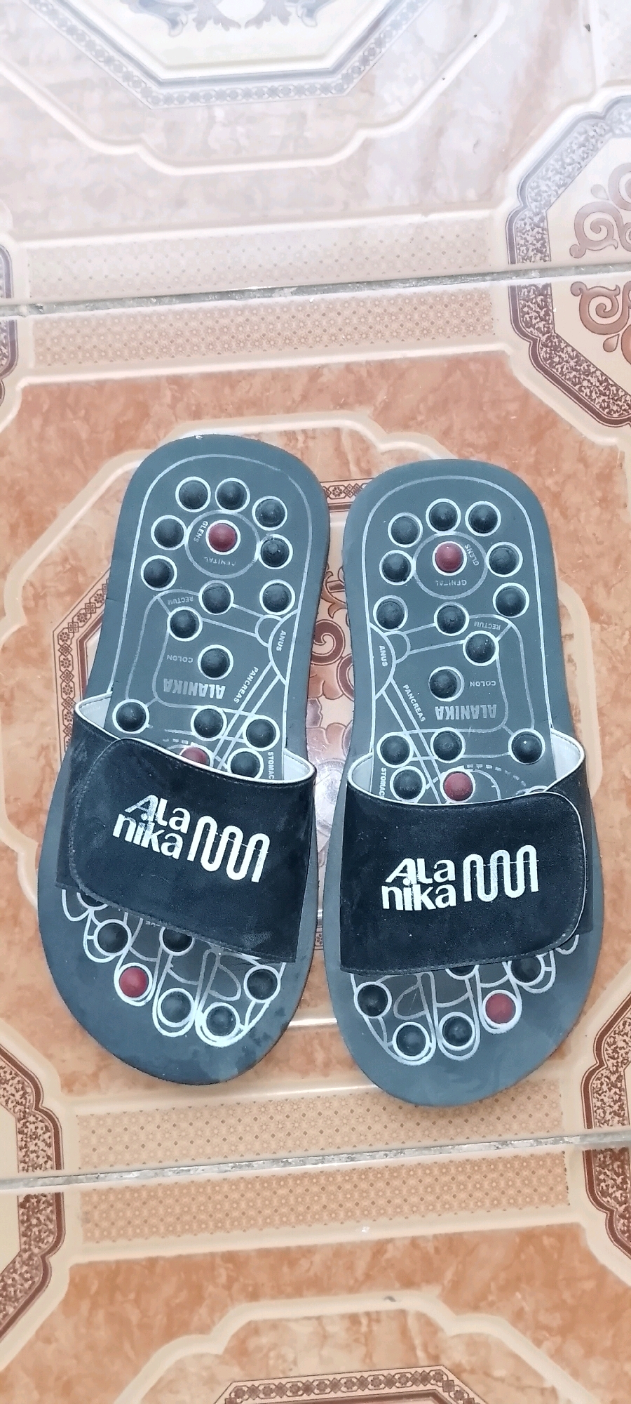 A pair of black, acupressure sandals lies flat on a patterned tiled floor. The sandals are designed with raised circular nodes and red dots, strategically placed to correspond to different parts of the foot, with labels such as "Colon," "Rectum," "Anus," and "Pancreas" visible on the footbeds. A black strap with a white logo that reads "Ala nika" crosses over the top of each sandal. The floor appears to be made of terracotta-colored tiles with decorative borders. The image is taken from a top-down perspective. The location is Satriamekar, Indonesia.